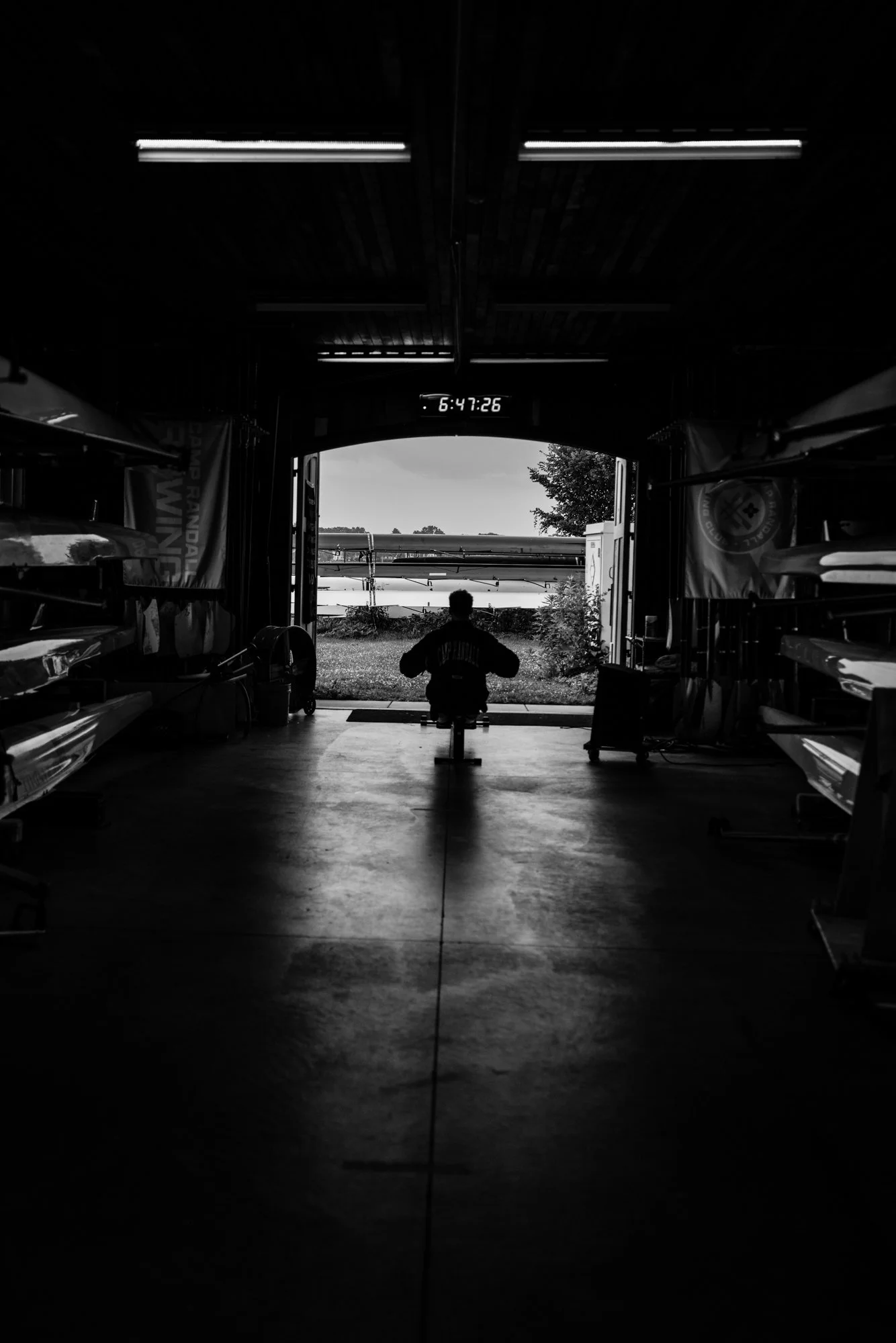 A person sitting on a bench inside a boat storage or workshop with an open garage door revealing an outdoor scene with water and trees, black and white photo.