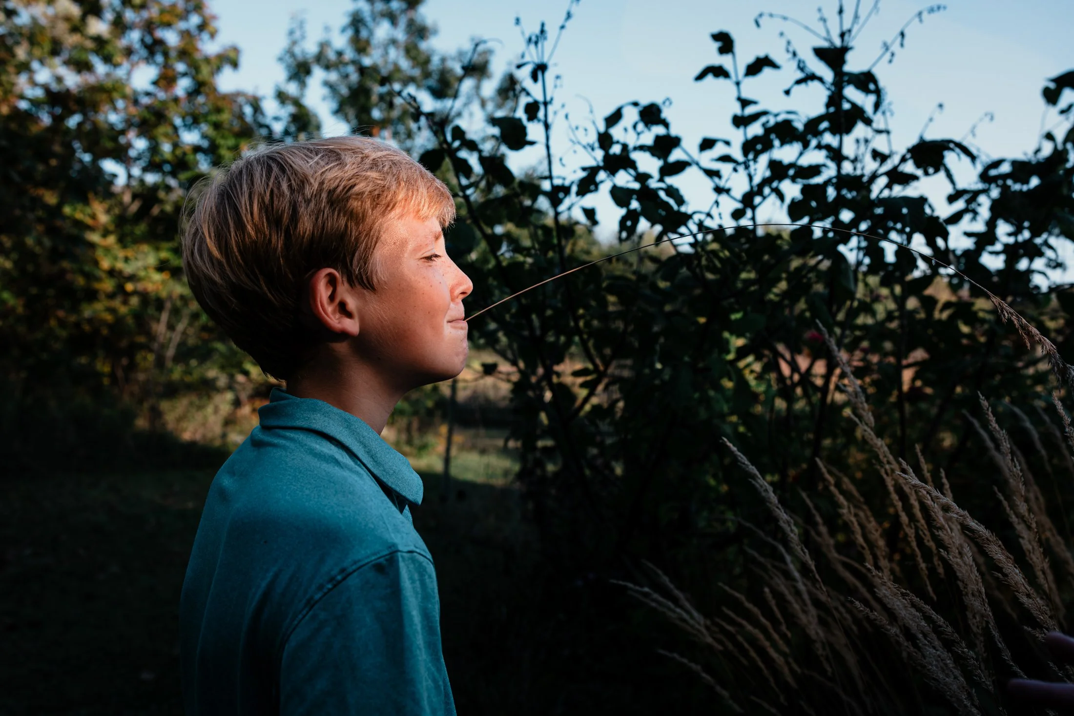 A young boy with short blonde hair and a teal shirt standing outdoors with eyes closed, enjoying the sunlight and nature around him.