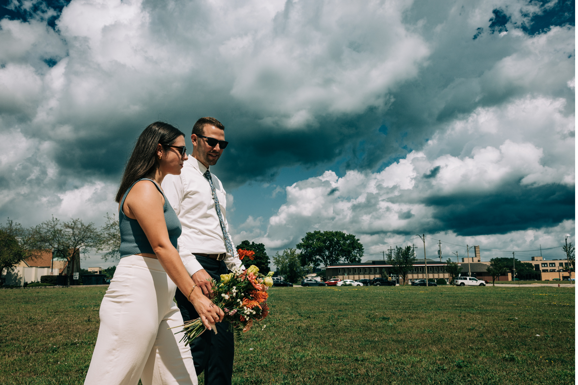 A woman and a man holding a bouquet of flowers walk on a grassy field under a cloudy sky.