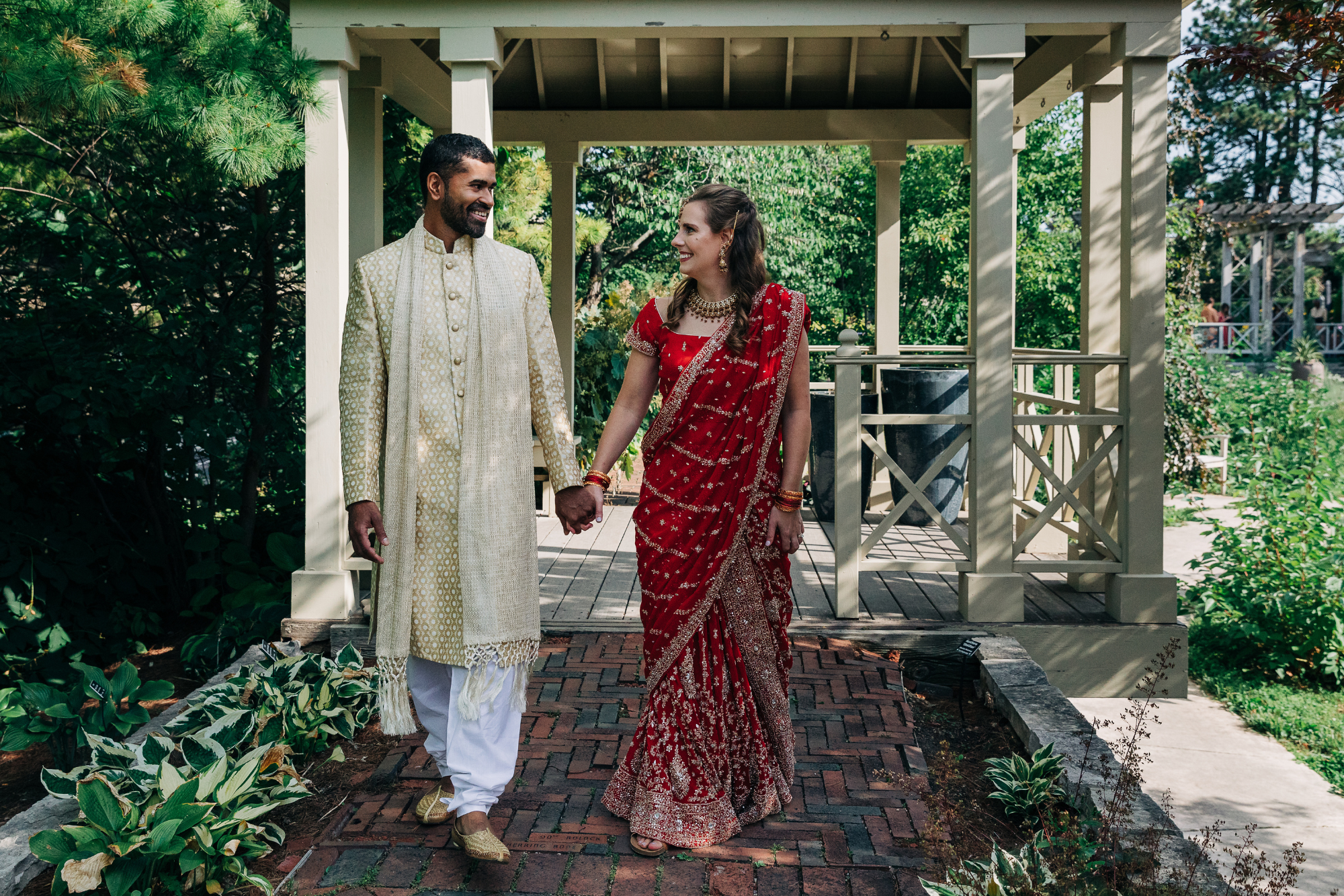 A couple in traditional Indian wedding attire holding hands and smiling at each other outdoors under a gazebo surrounded by greenery.
