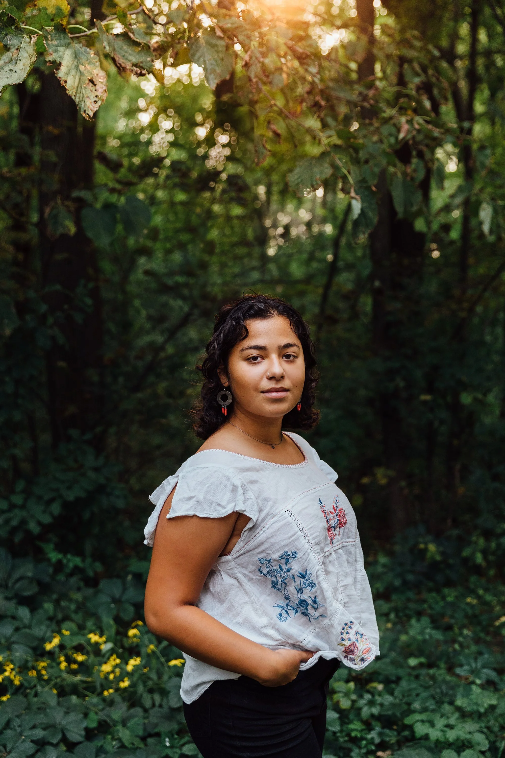 A young woman with curly dark hair, wearing earrings and a white embroidered blouse, standing outdoors in a forested area with sunlight filtering through the trees.