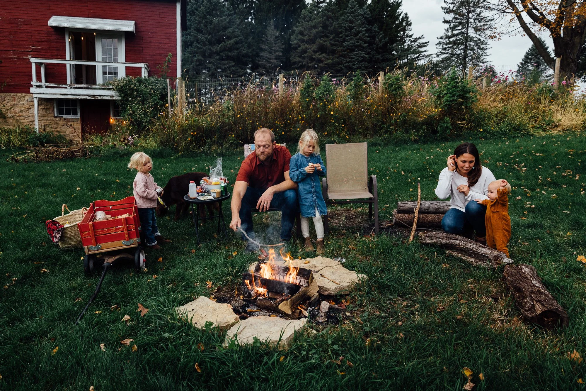 A family gathering around a small campfire in a backyard during fall. A man is tending the fire while two young children sit nearby, one on a chair and the other standing. Another woman is sitting on logs, holding a smiling baby. The yard features a 