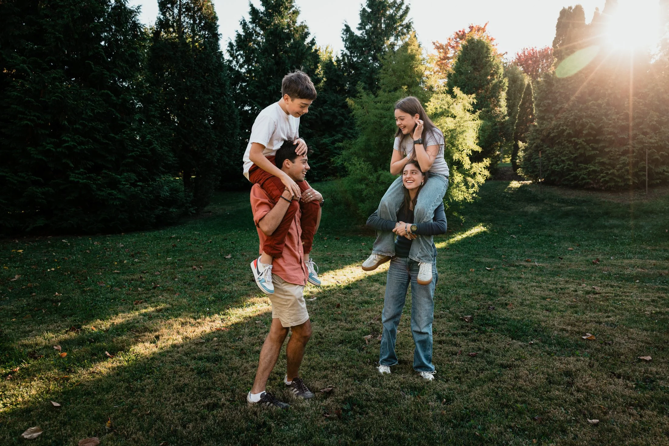 Group of four young friends in a park, with two men and two women, sharing a joyful moment as the person on the right is giving the person on the left a piggyback ride near trees and sunlight.