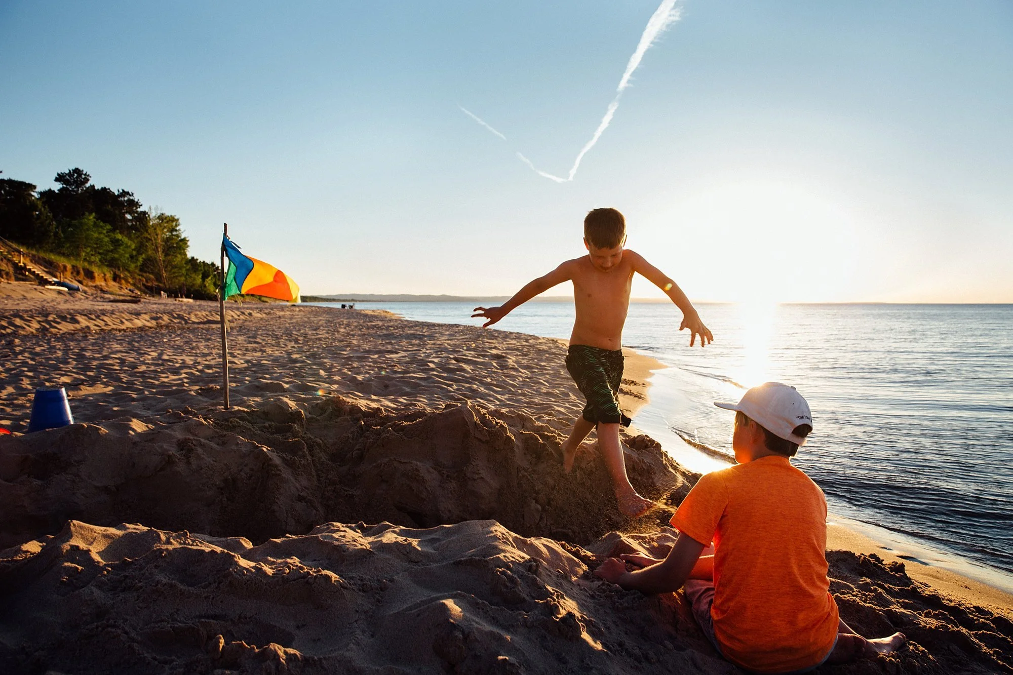 Two boys playing on a beach at sunset, one is jumping into a sandpit while the other sits nearby watching, with a colorful umbrella and a blue cup placed on the sand.