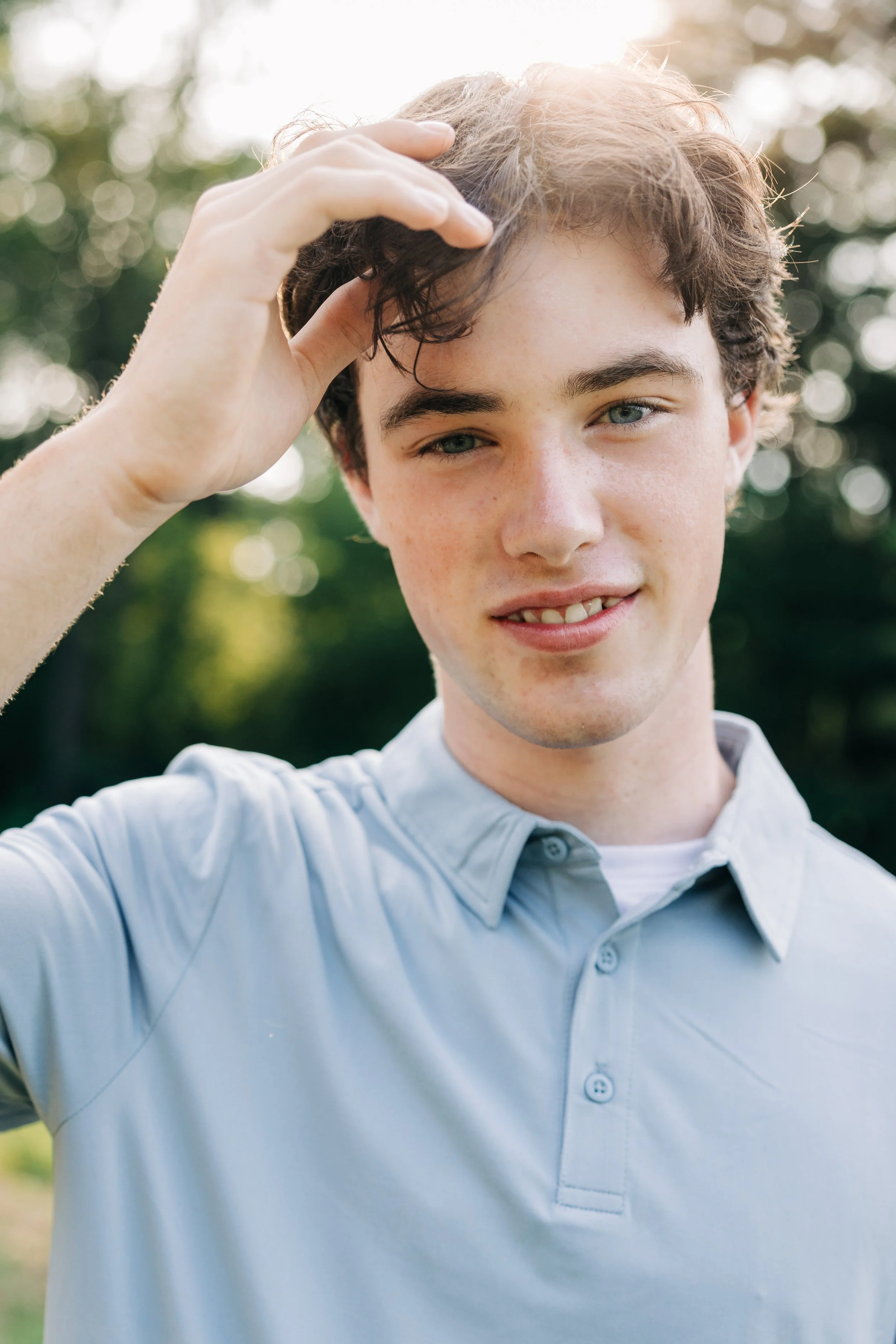 A young man with curly brown hair and freckles, wearing a light gray polo shirt, is outdoors with greenery in the background, touching his hair and smiling at the camera.