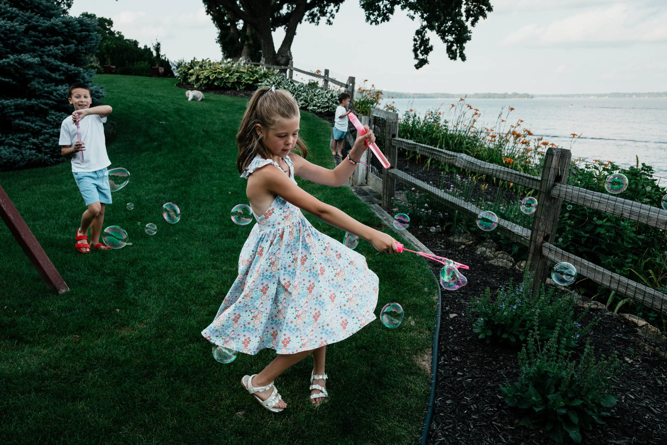 Children playing with bubbles near a lake, with a girl in a floral dress blowing bubbles and a boy in a white shirt and blue shorts running.