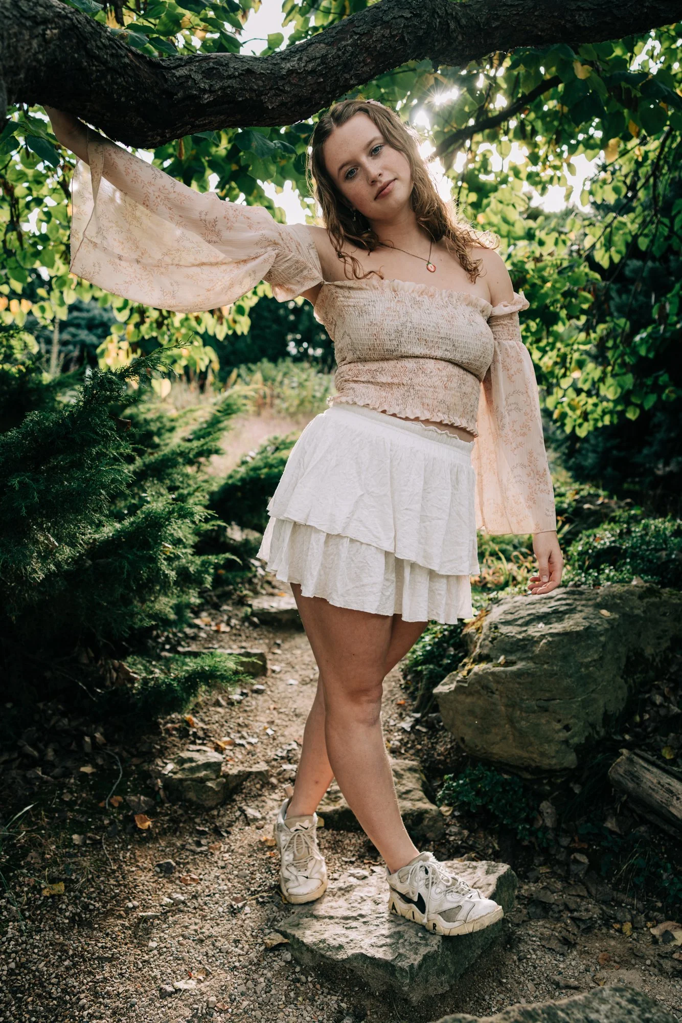 Young woman in a beige off-shoulder top and white tiered skirt standing on a stone in a lush green garden or forest, with sunlight filtering through the trees.