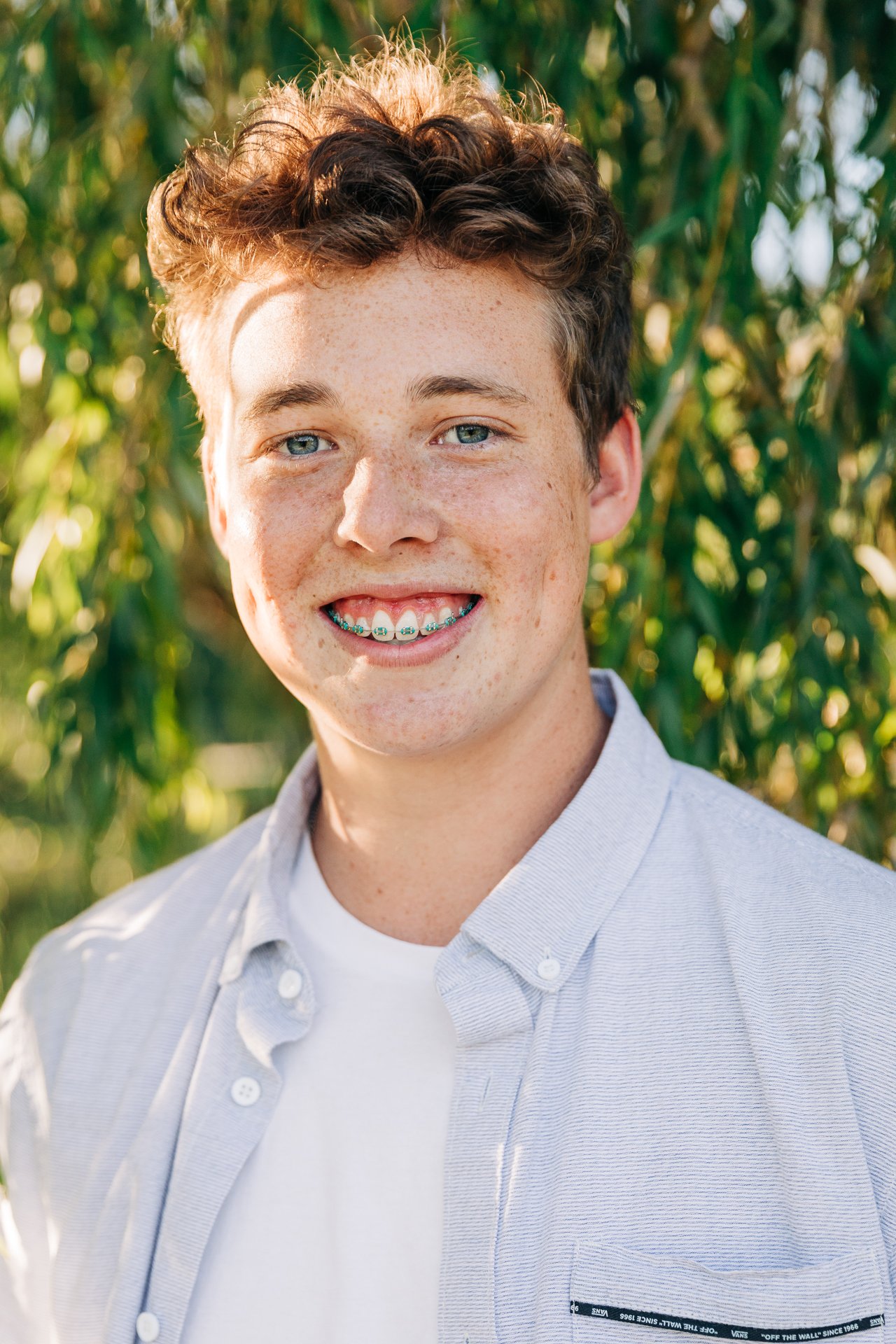 A teen boy with curly brown hair and freckles, smiling, showing braces, wearing a light gray button-up shirt and white t-shirt, outdoors with green foliage in the background.