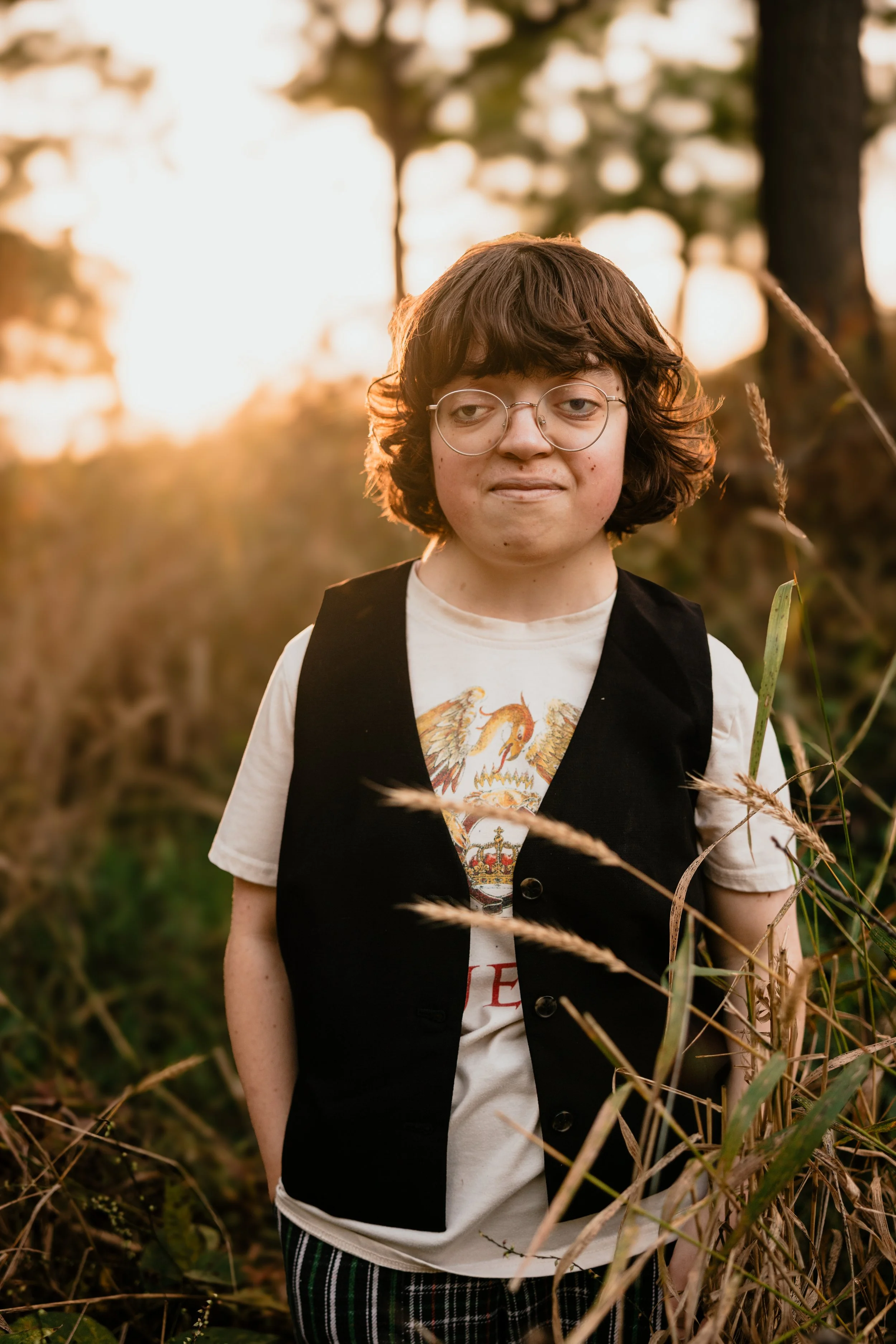 A young person with curly brown hair and glasses standing outdoors among tall grass during sunset, wearing a white t-shirt with a royal crest and a black vest.