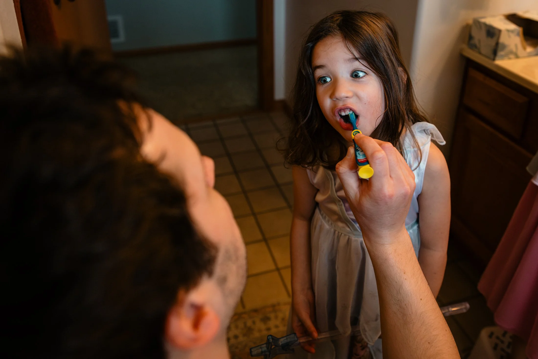 A young girl with brown hair and blue eyes brushing her teeth while an adult helps her by holding the toothbrush, inside a home with a wooden cabinet and tiled floor.