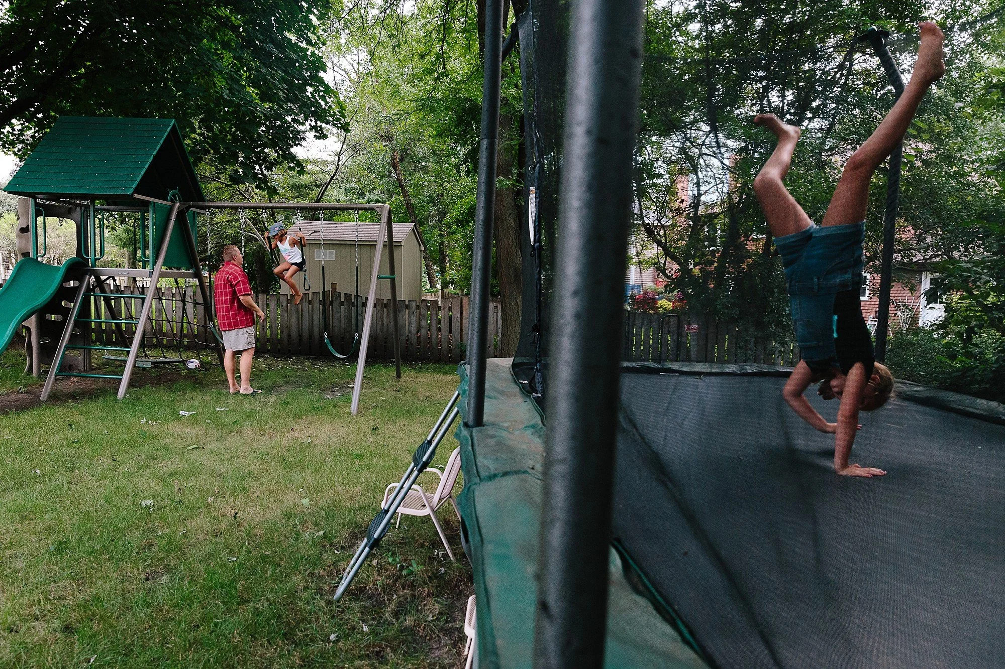 Child upside down on a trampoline and two kids playing on a swing set in a backyard with trees and a fence.