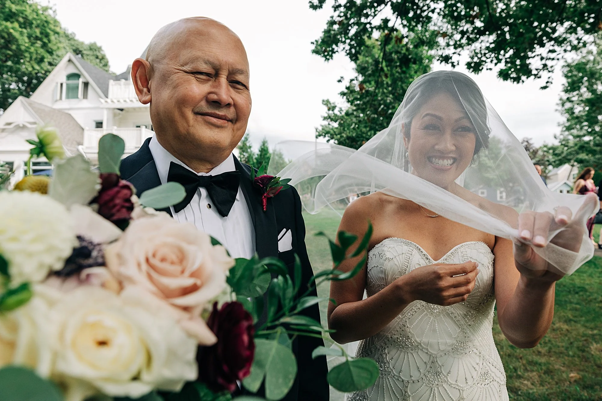A bride and an older man on her wedding day outdoors, the bride smiling and holding her veil, the man smiling with closed eyes, a bouquet of flowers in the foreground, manicured lawn and a white house with gabled roofs in the background.