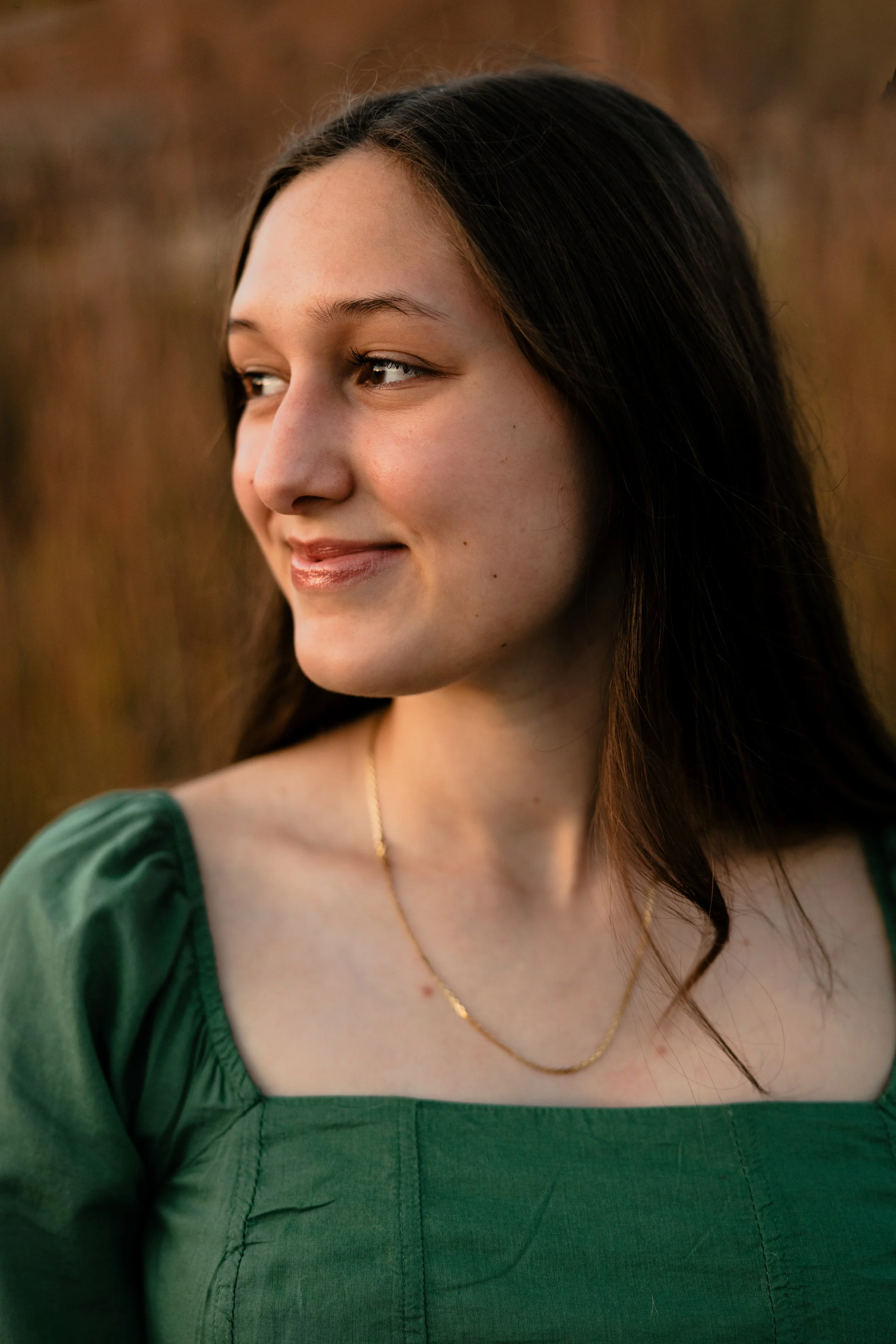 A young woman with long dark hair smiling softly, wearing a green top and a gold necklace, outdoors during sunset.