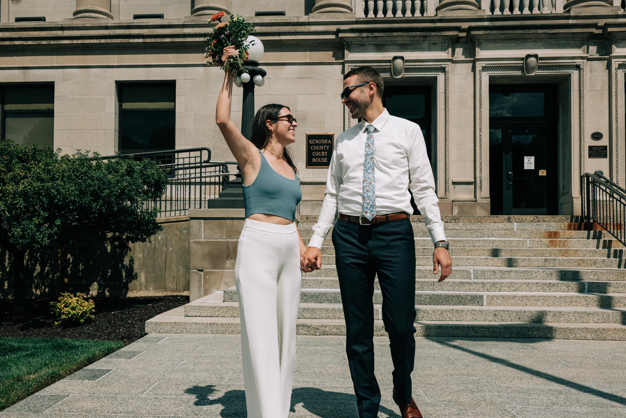 A couple holding hands outside a courthouse, smiling and celebrating, with one woman holding a bouquet of flowers in the air.