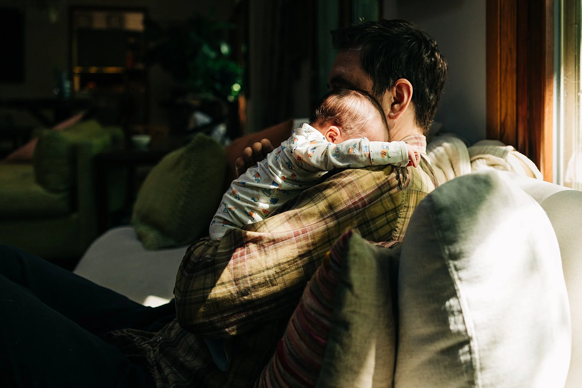 A man sitting on a beige couch with a green pillow, holding a sleeping baby on his shoulder in a dimly lit living room, with sunlight coming through a window.