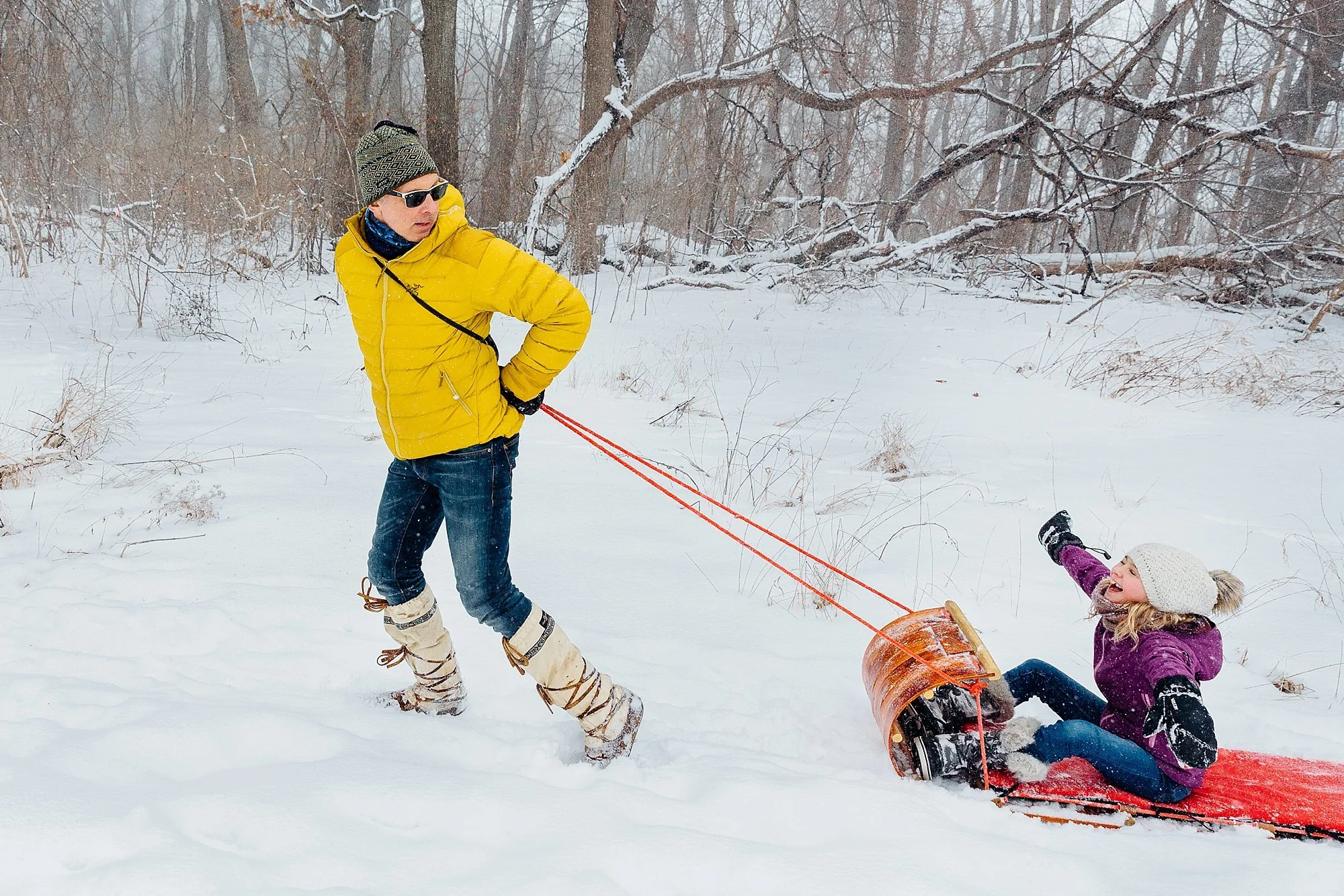 A man in a yellow jacket and jeans pulls on a rope attached to a child sitting on a red sled in a snowy forested area.