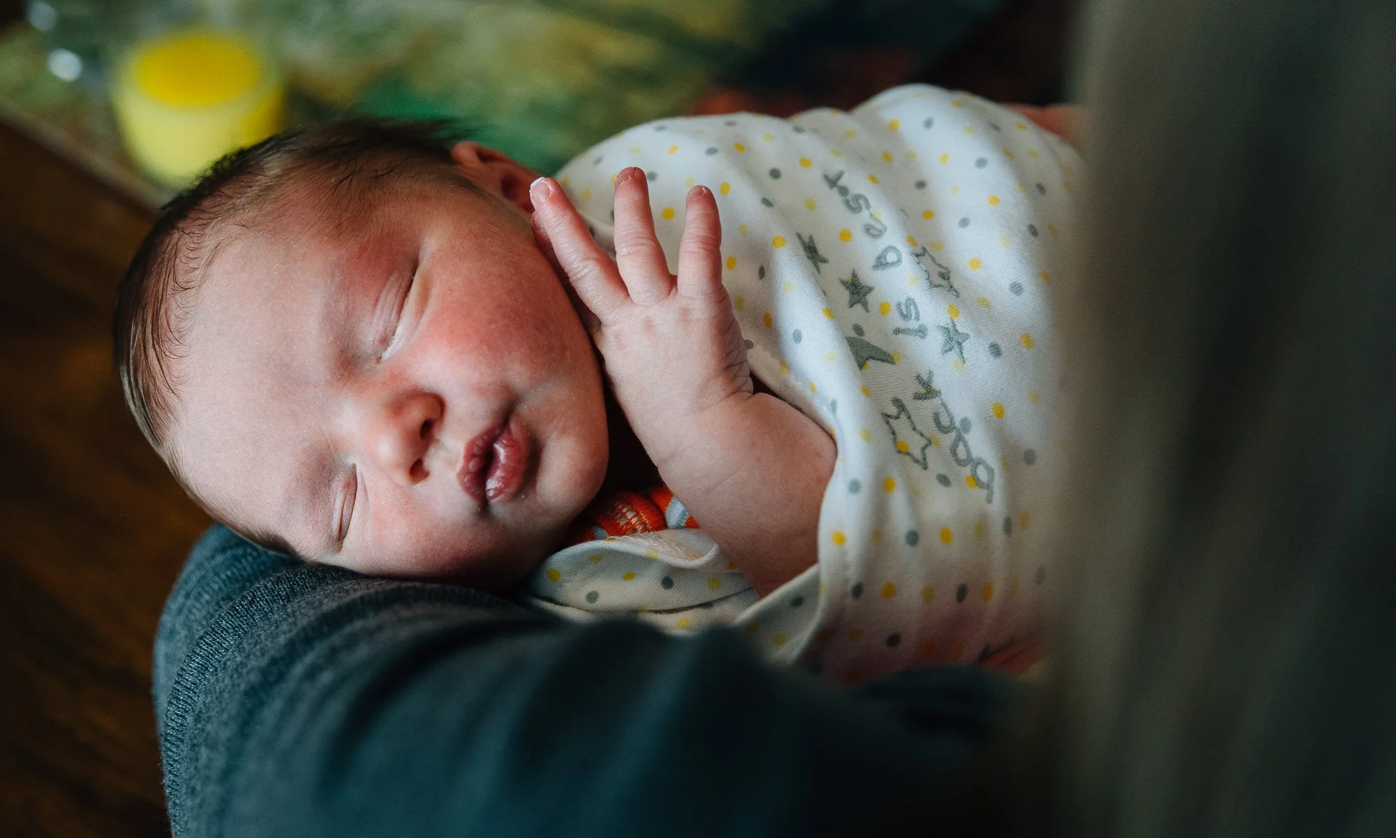 A newborn baby peacefully sleeping and resting on a person's chest, with one hand near their face, wearing a white outfit with colorful polka dots and star patterns.