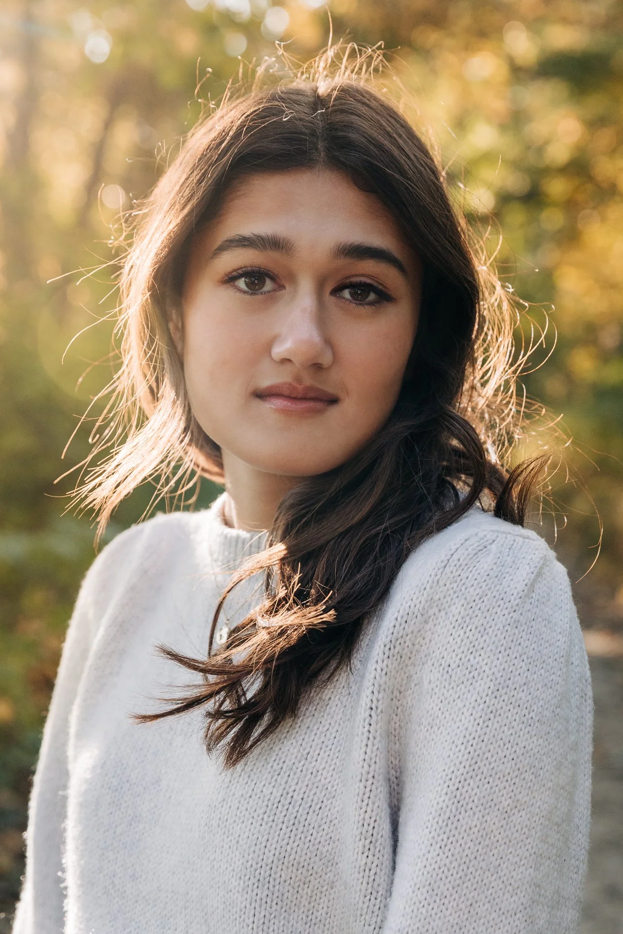 Portrait of a young woman with long dark hair wearing a white knitted sweater outdoors in a natural setting during golden hour.