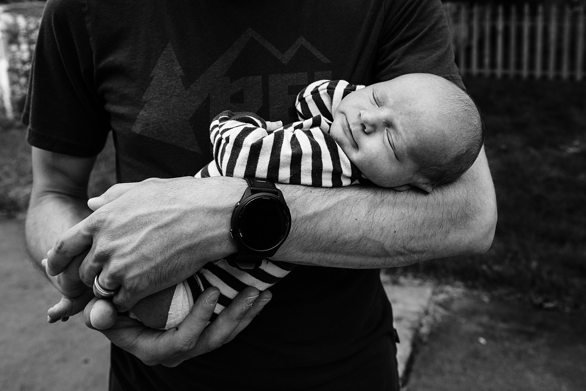 A person holds a sleeping baby in their arms outdoors, with a fence in the background. The baby is wearing a striped outfit.