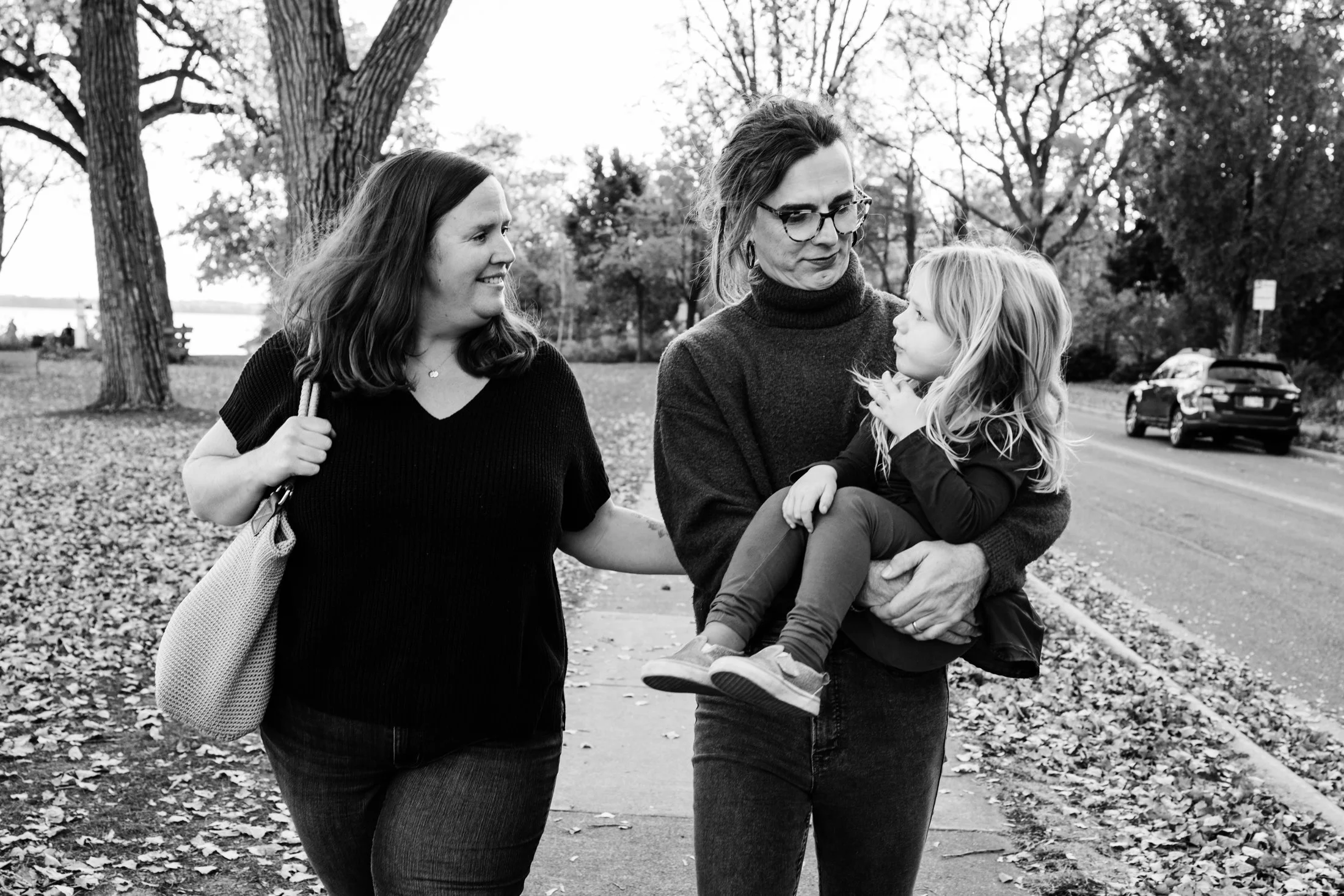 Three women, one carrying a young girl, walking outdoors in a park with trees and fallen leaves.