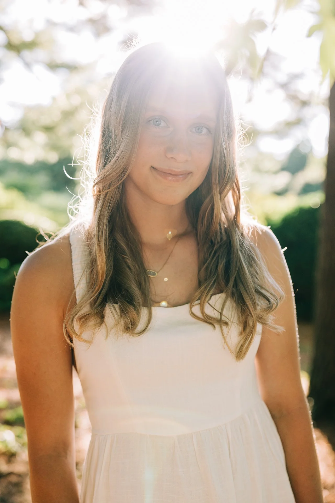 A young woman with long, wavy hair stands outdoors in sunlight, wearing a white dress and layered necklaces, with trees and greenery in the background.