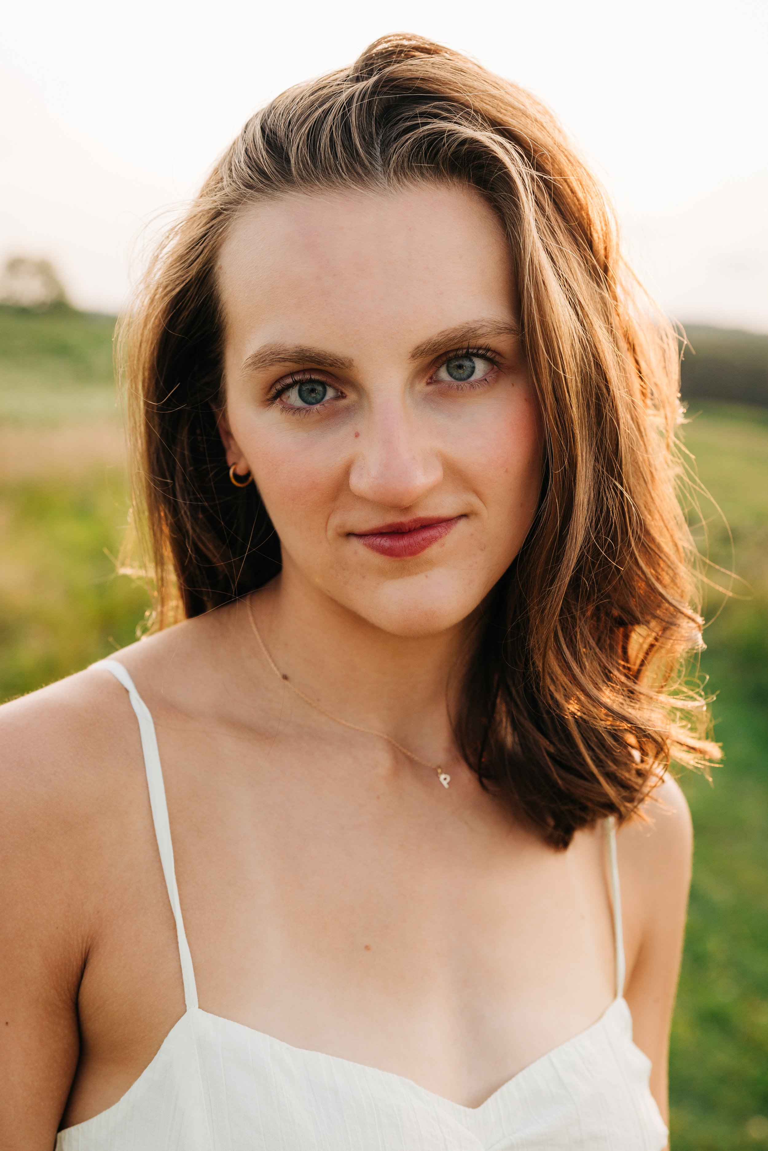 Close-up of a young woman with wavy brown hair, blue eyes, and light skin, wearing a white spaghetti strap top, standing outdoors in a grassy area during golden hour with soft sunlight