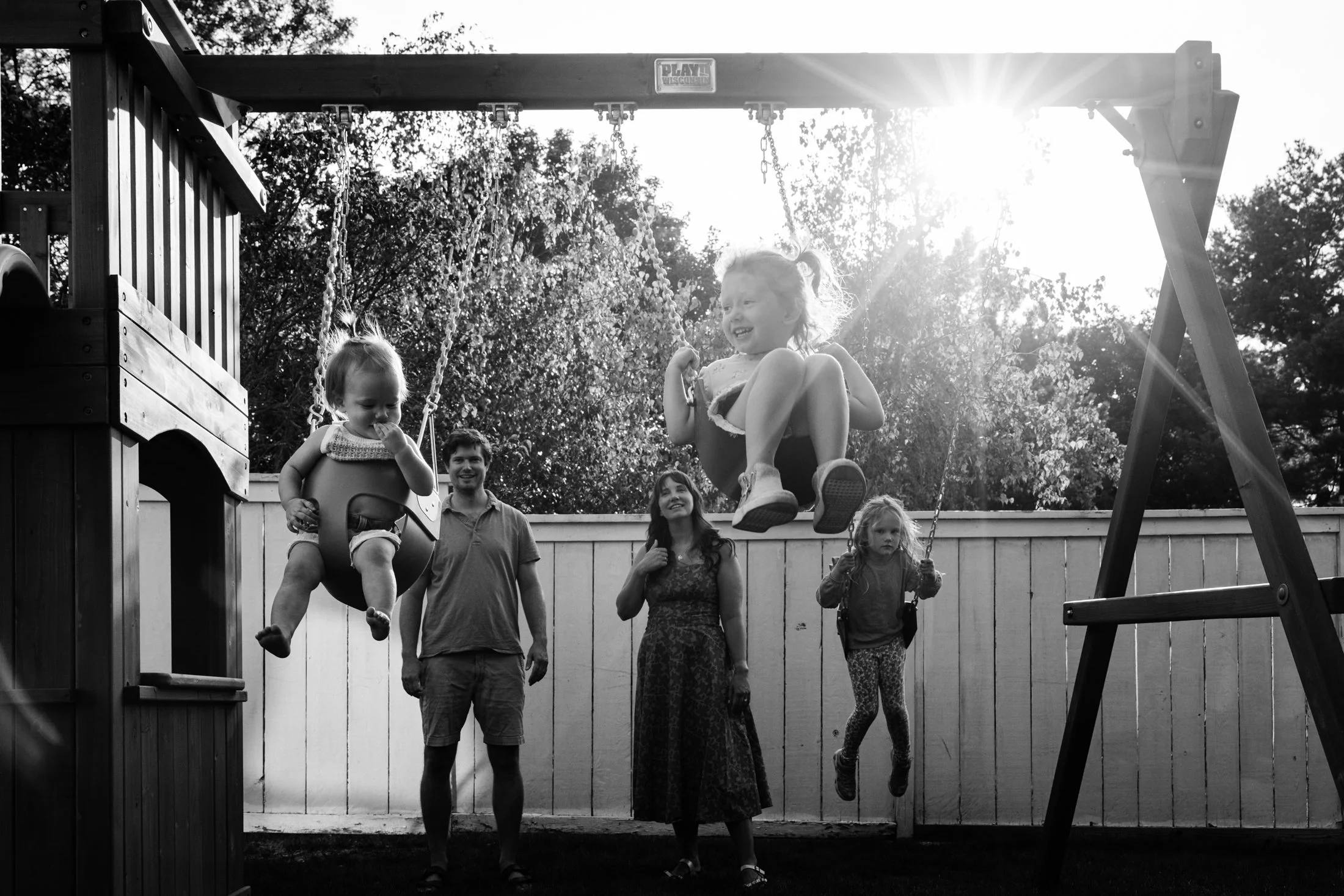 A black and white photo of children playing on a swingset in a backyard with a fence and trees in the background. Two kids are swinging on the swings, one girl is in a bucket swing on the left, and another girl is in a traditional swing on the right.