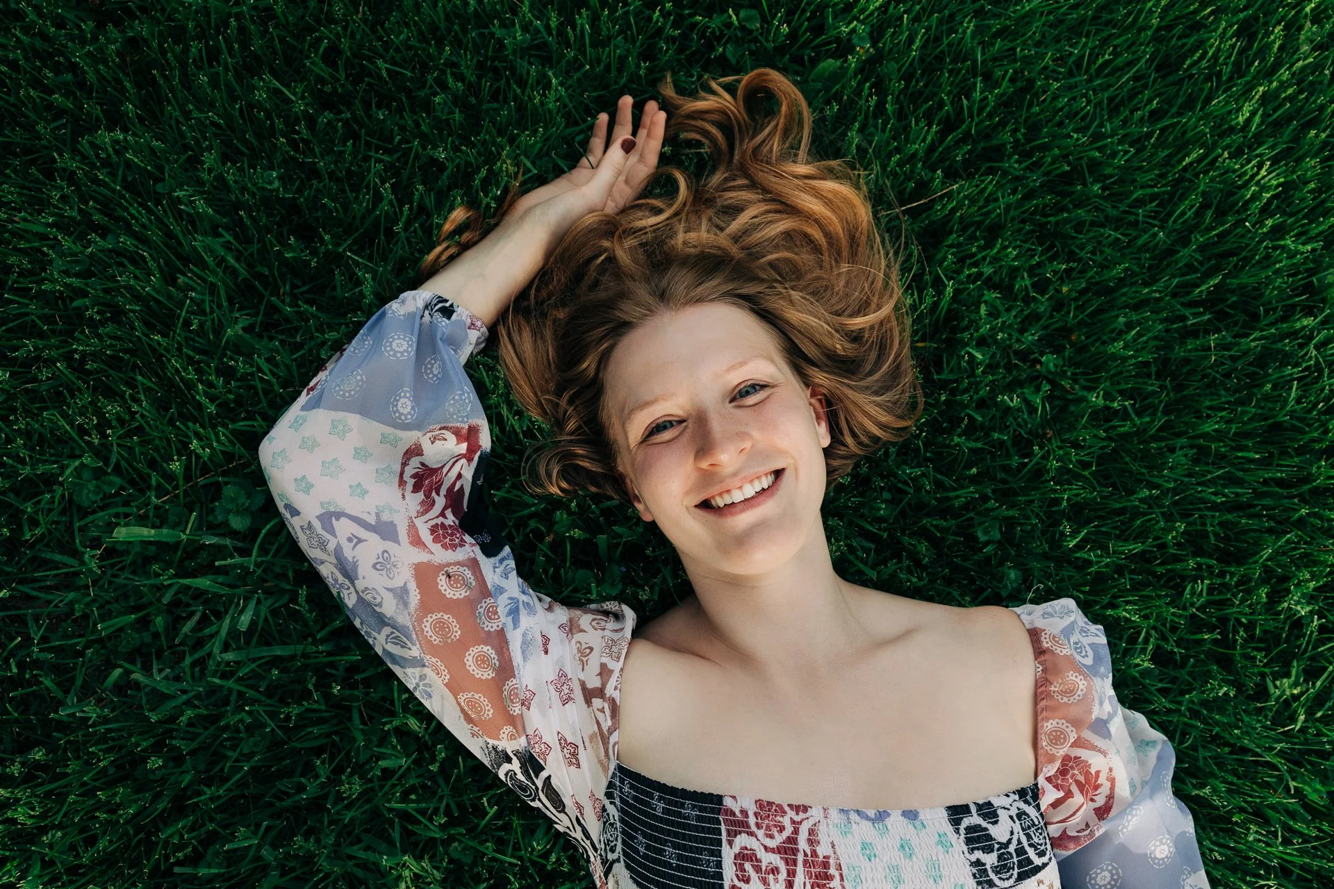 A young woman with curly red hair lying on green grass, smiling at the camera.
