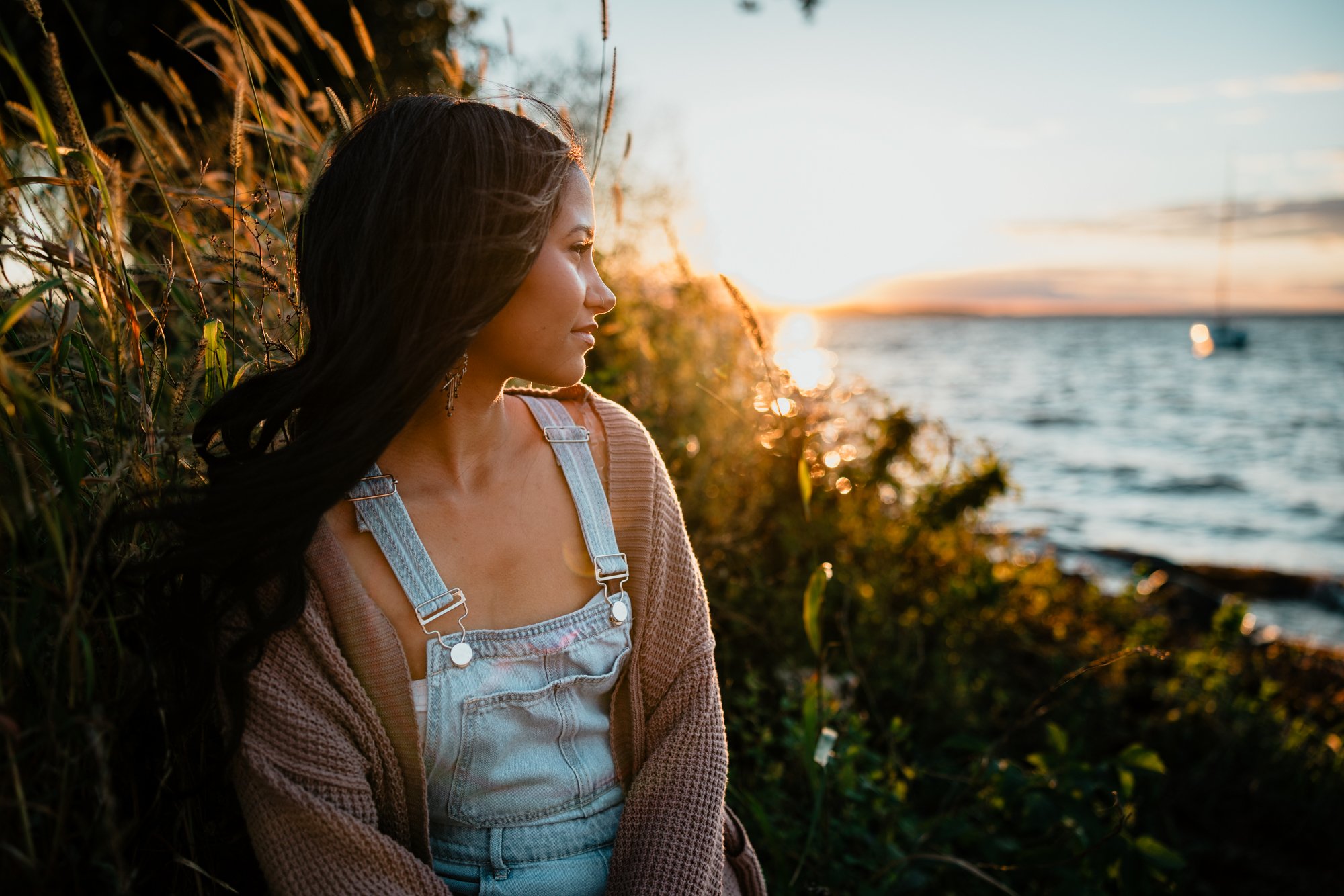 A woman sitting beside bushes by a body of water during sunset.