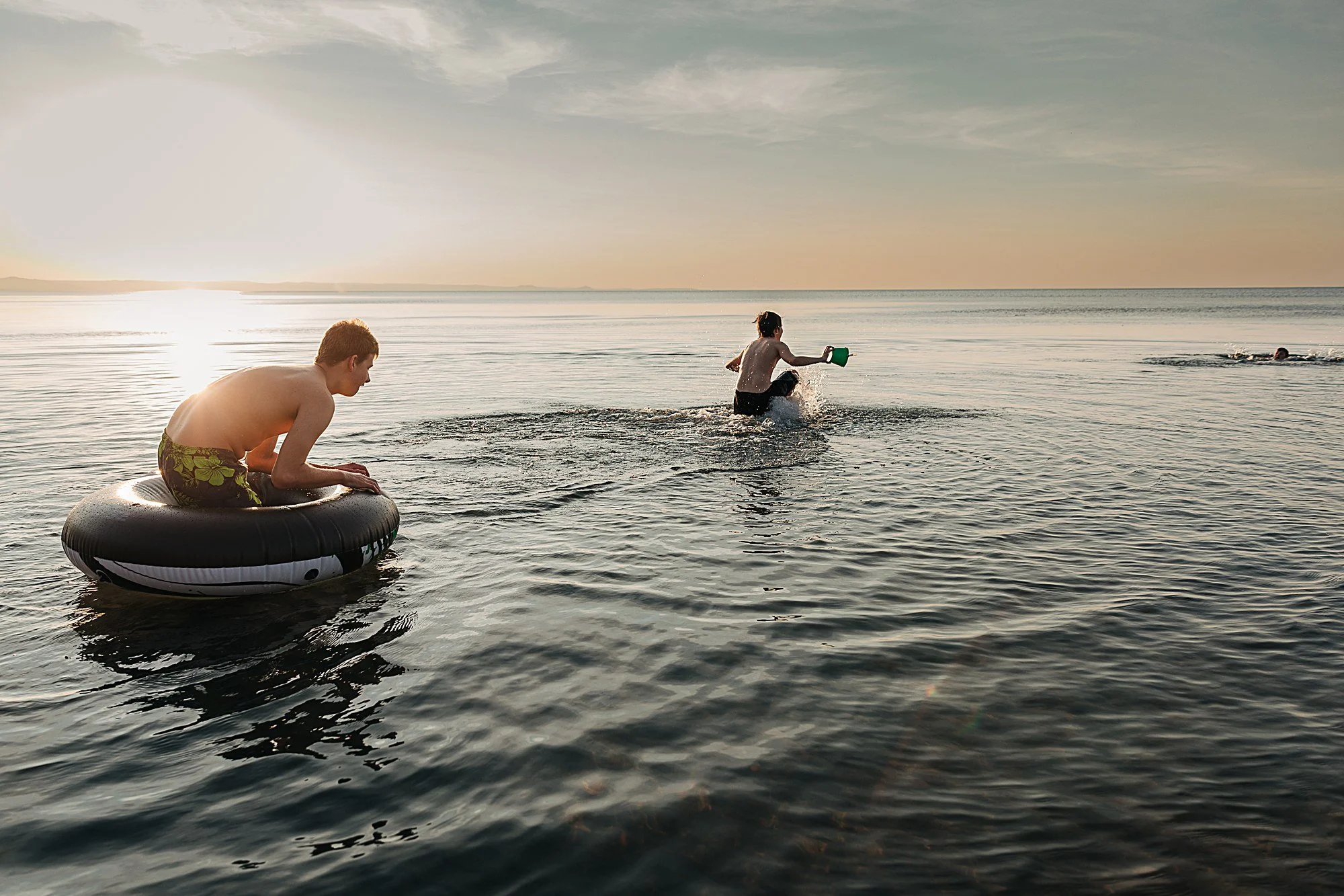 Three children playing in the water at the beach during sunset. One child is sitting on an inflatable ring, another is running with a plastic bucket, and a third is swimming in the distance.
