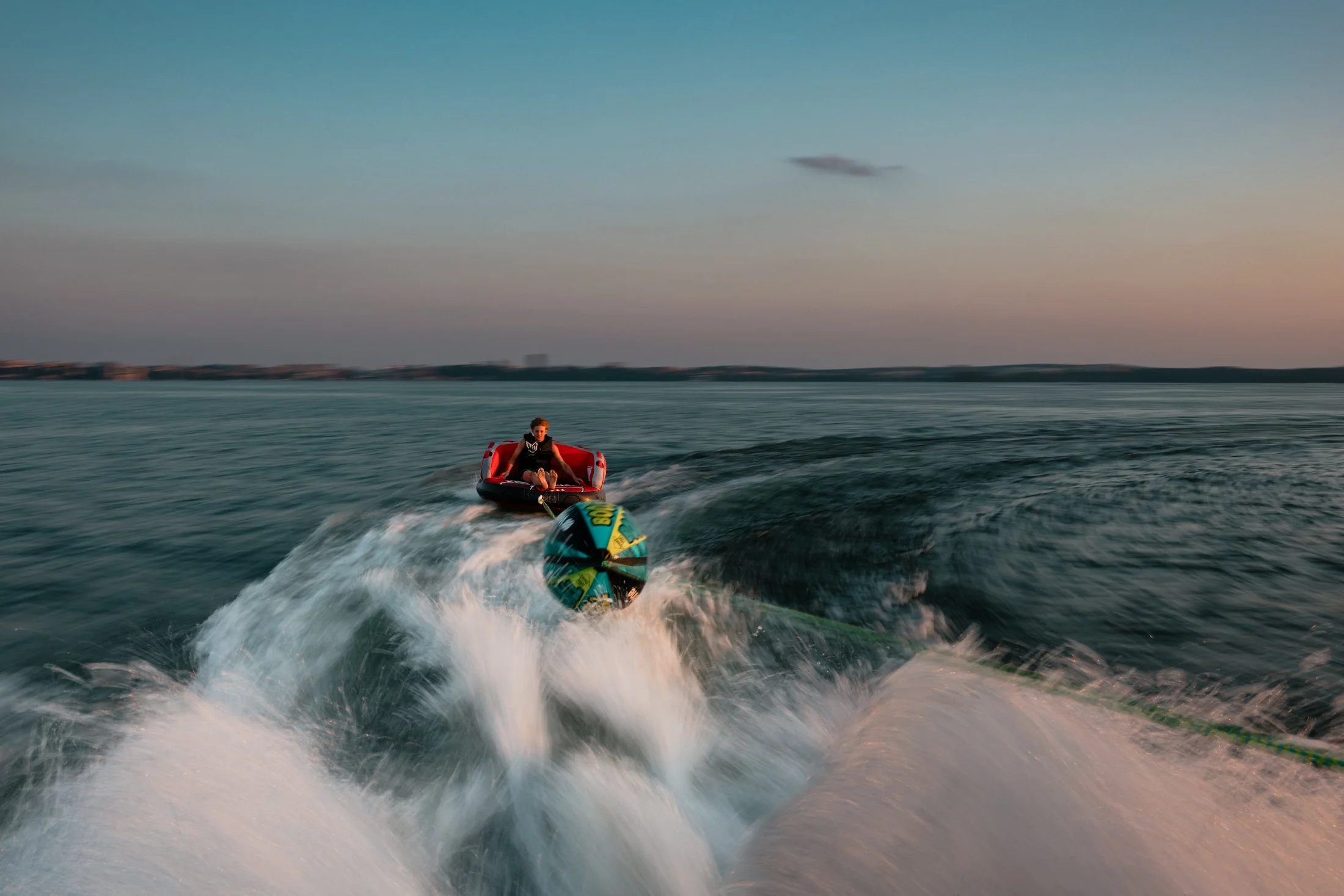 Person riding a watercraft pulled by a jet ski on a large body of water during sunset.