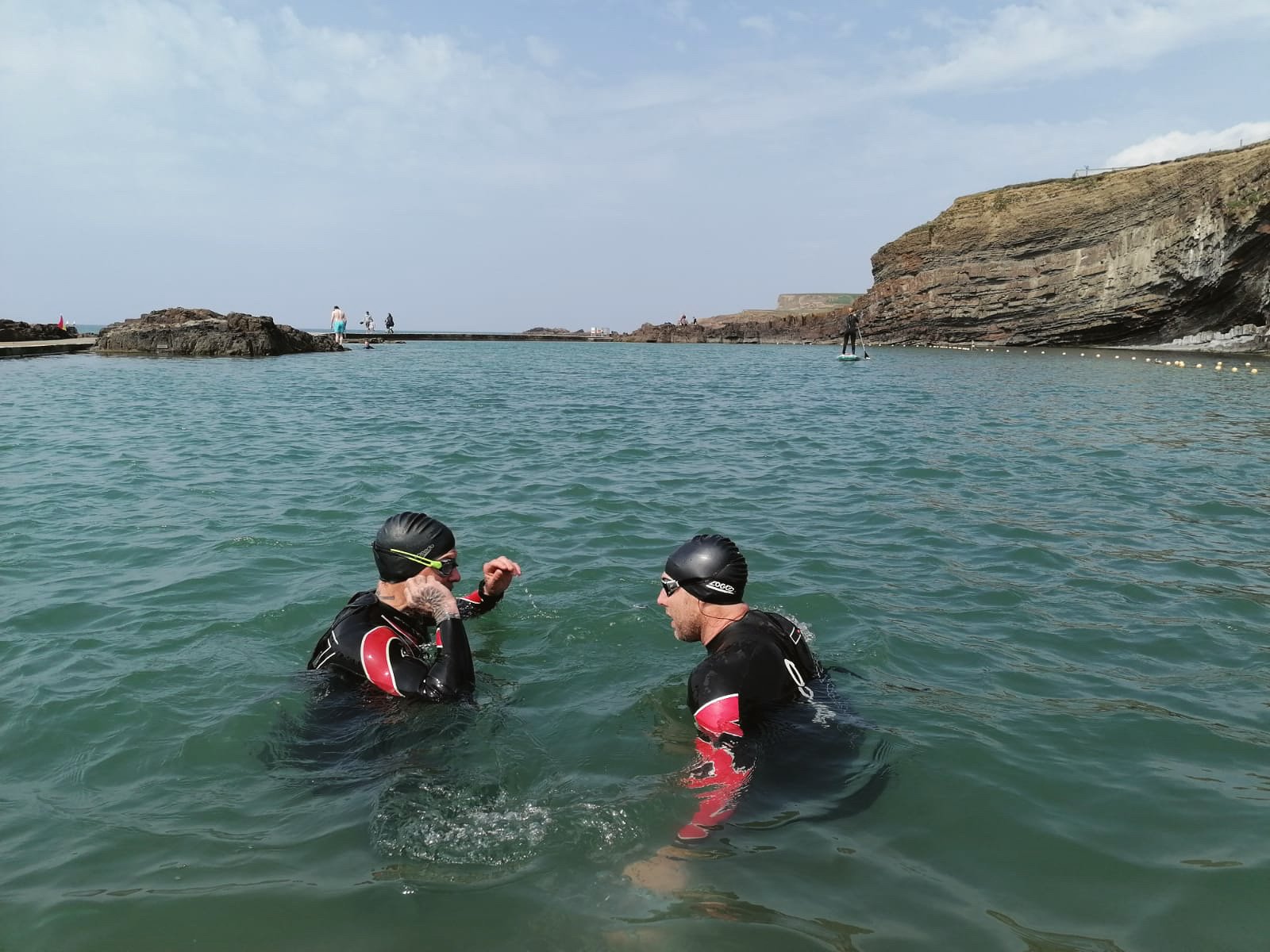 Two people wearing black wetsuits and helmets standing in the ocean, engaging in conversation during a sunny day near rocky cliffs.