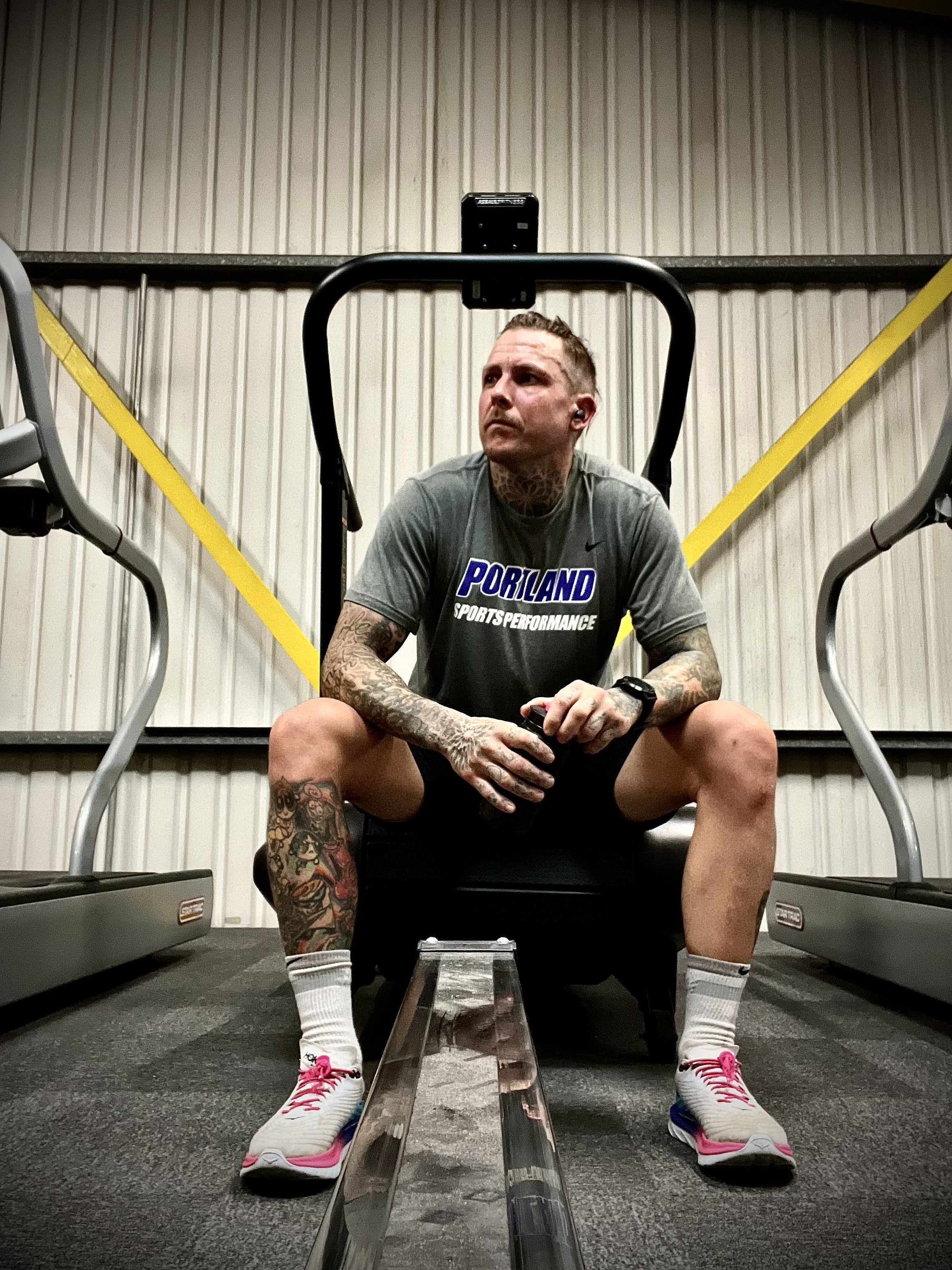 A man with tattoos on his arms and legs, wearing a gray Portland sports performance t-shirt, sitting on a treadmill in a gym, looking to the side, holding a small water bottle, with a phone mounted overhead.
