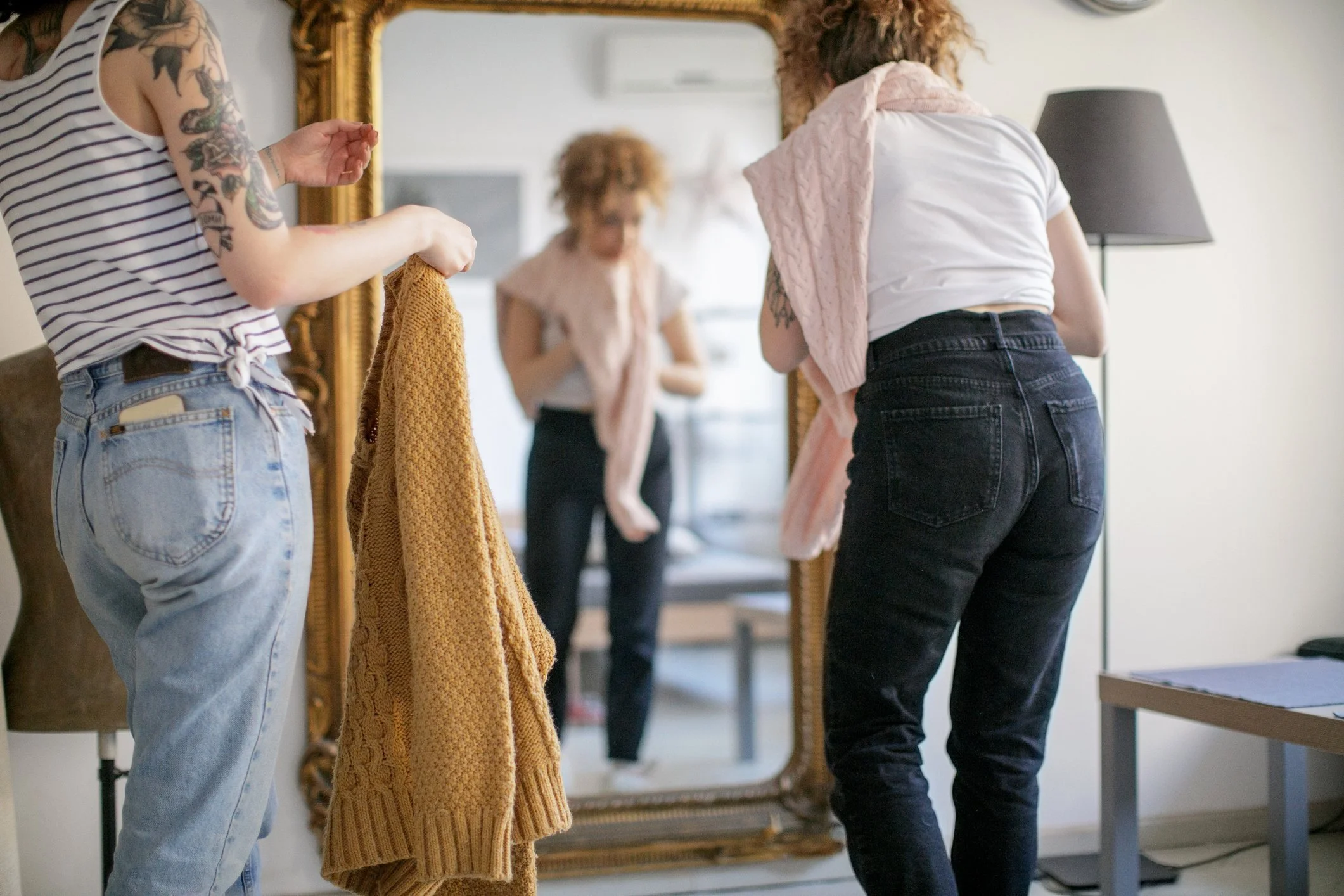 Two people in casual clothing are trying on sweaters in front of a large ornate mirror in a home setting. One person is wearing a striped tank top and holding a mustard-colored sweater, while the other has a light pink sweater draped over their shoulders.