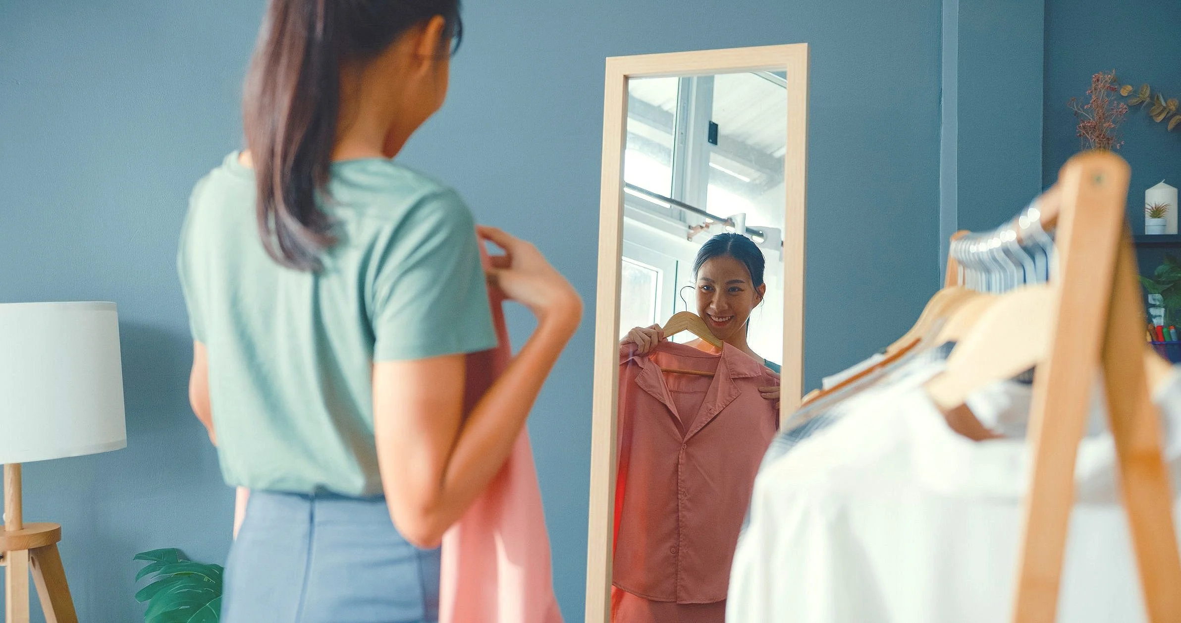Woman trying on clothes in front of a mirror in a blue room with a clothing rack.