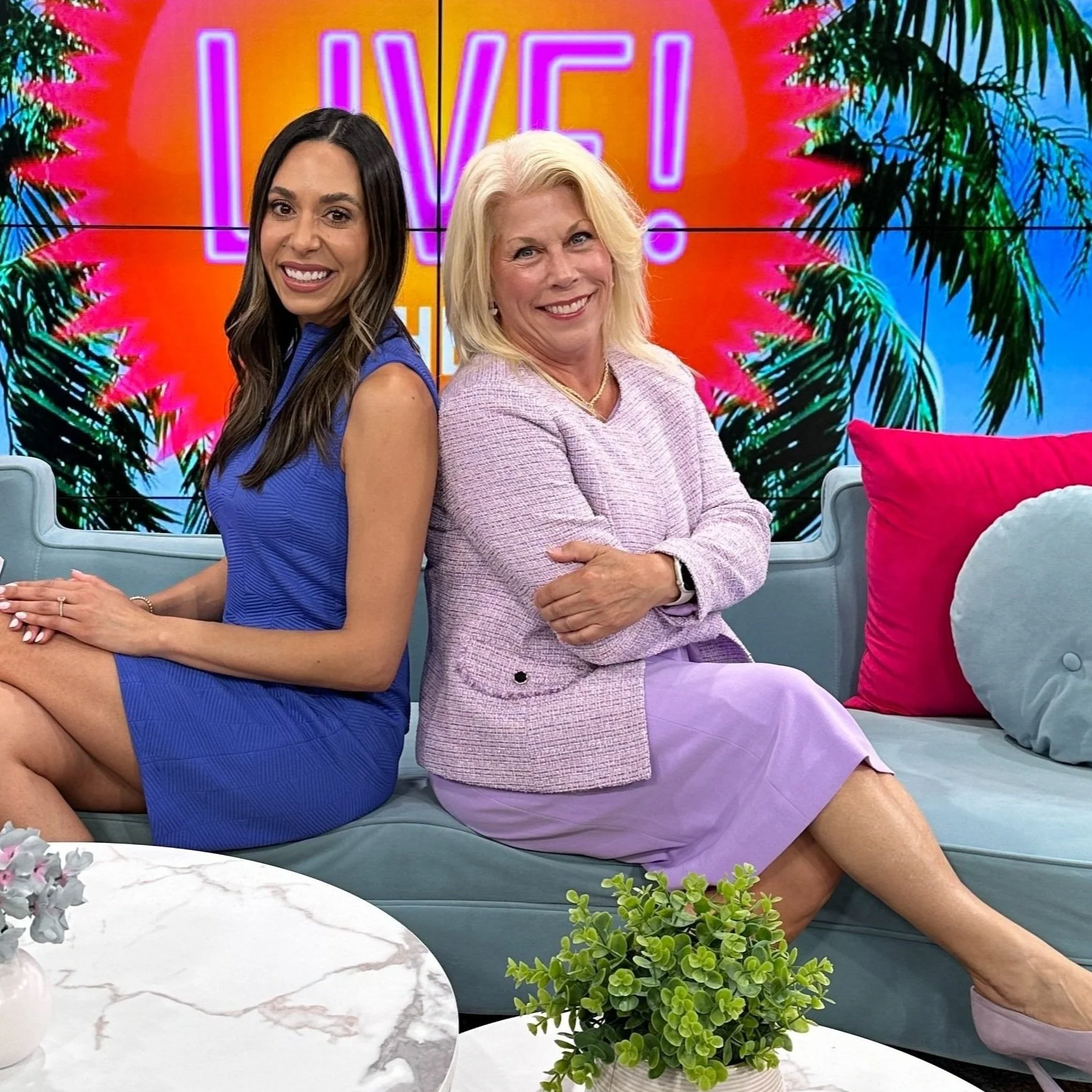 Two women sitting on a couch in a TV studio, smiling, with a colorful background and the word 'LIVE!' displayed. One woman is wearing a blue dress and the other a pink outfit. The setting includes vibrant décor with plants and colorful cushions.
