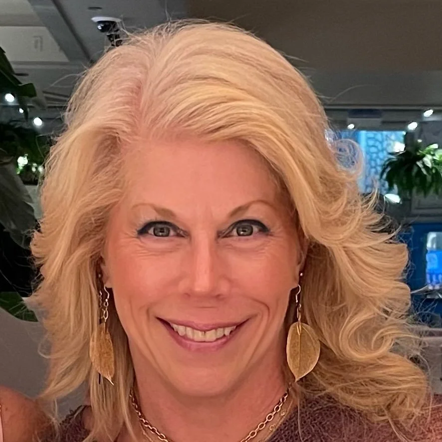 Smiling woman with wavy blonde hair wearing leaf-shaped earrings and a necklace, indoors with blurred background of plants and lights.