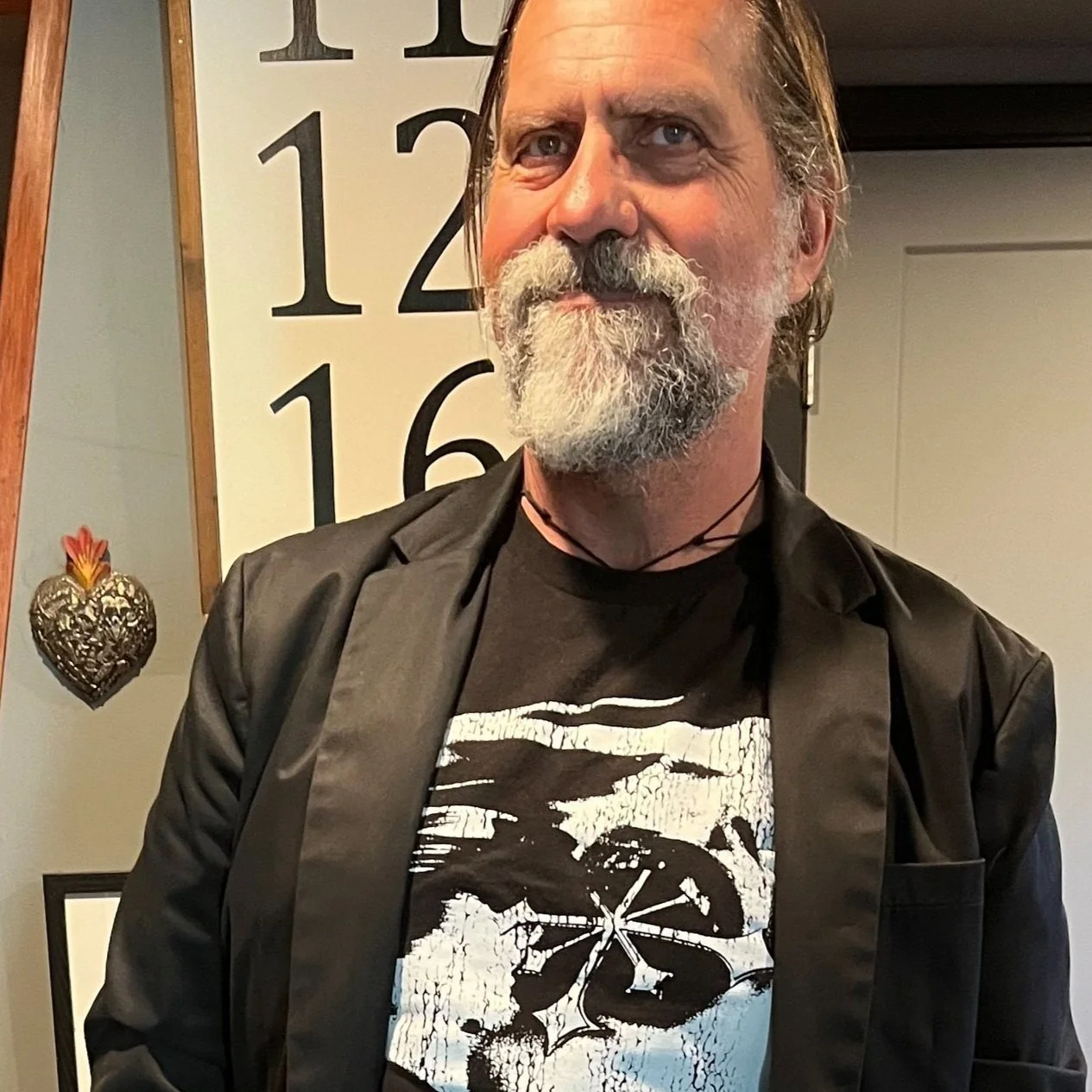 Man with a beard wearing a black jacket and graphic t-shirt, standing in front of a wall with numbers.