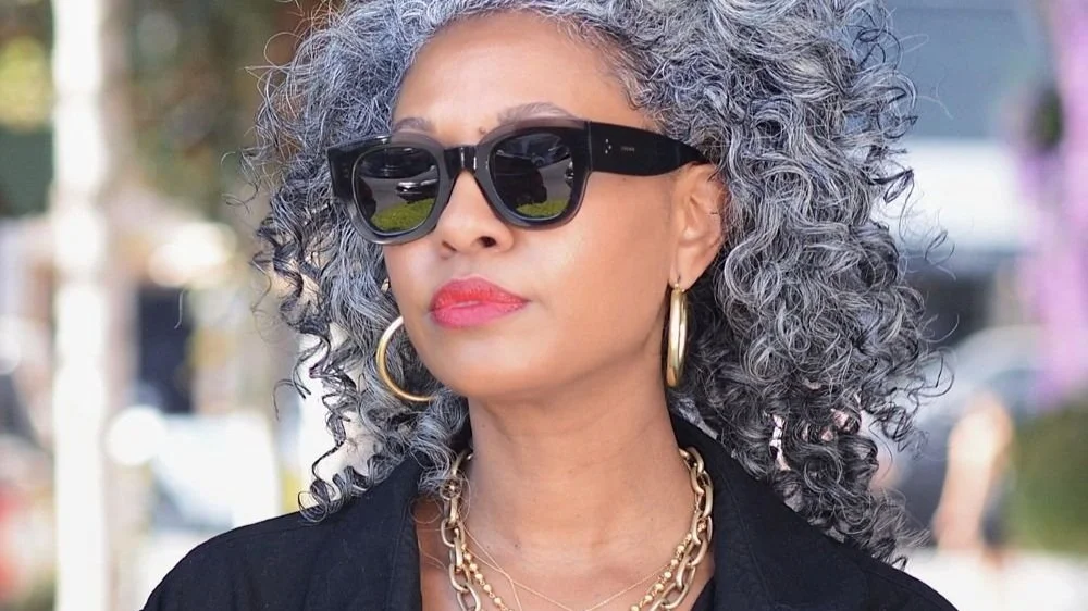 Stylish woman with curly gray hair wearing sunglasses and gold jewelry.