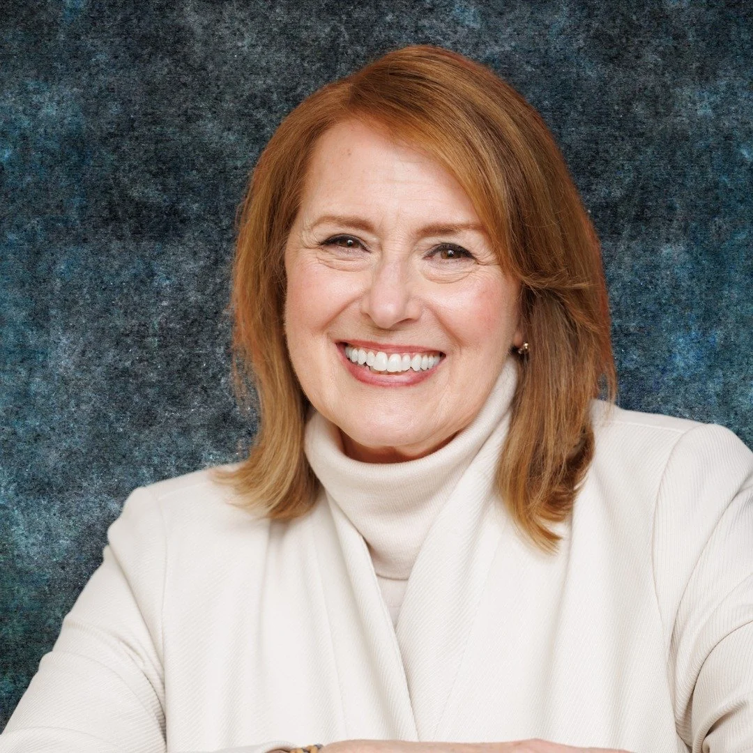 Smiling woman with red hair wearing a beige turtleneck and jacket against a blue textured background.