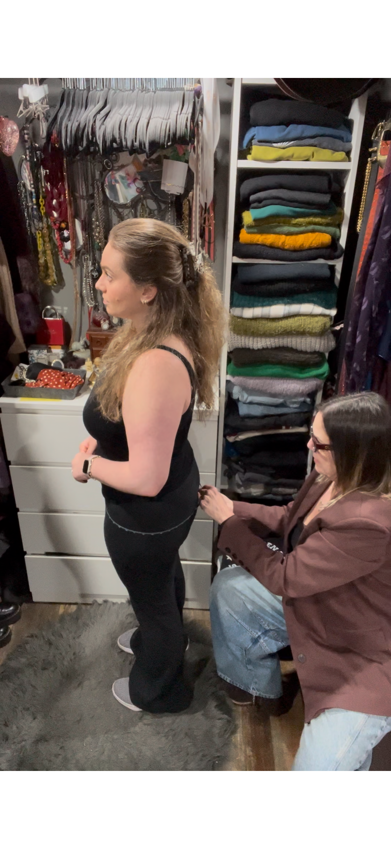 Two people in a closet; one standing, the other kneeling to adjust clothing.