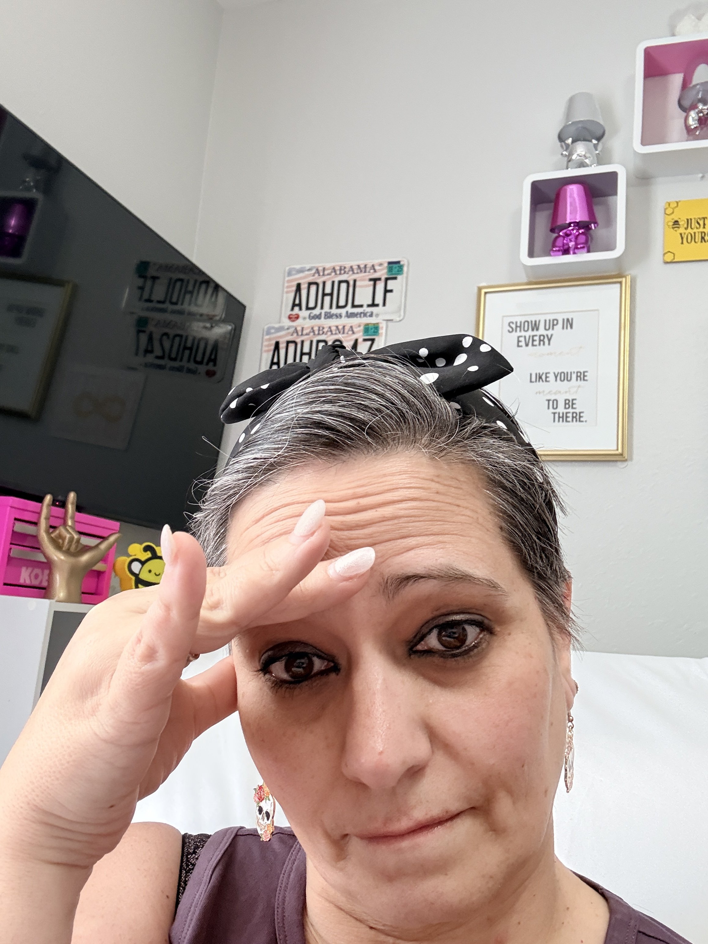 A woman with gray hair wearing a black headband with white polka dots, sitting on a bed or couch, with I can't believe it expression and her hand resting on her forehead.
