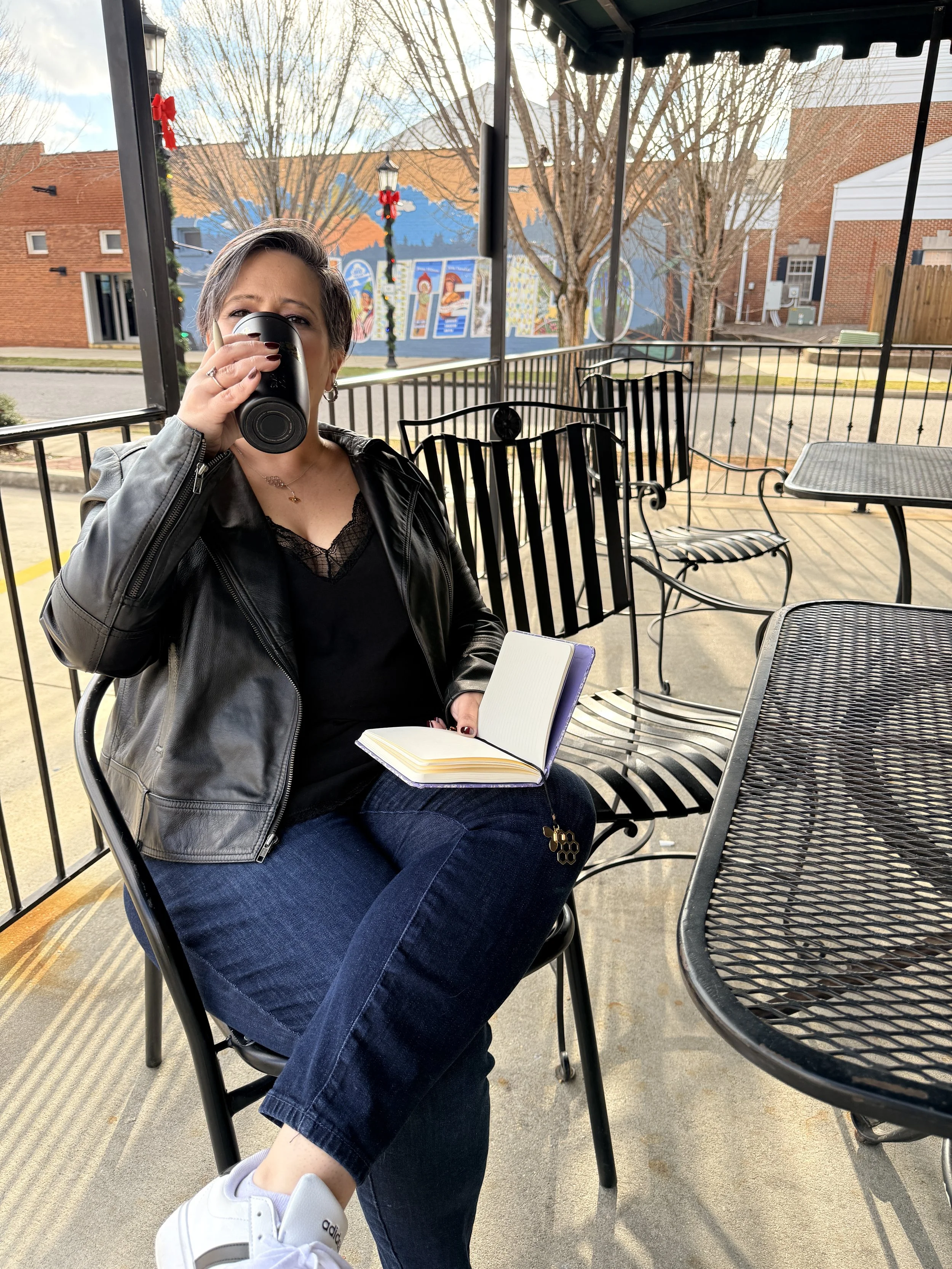 A woman sitting at an outdoor table, drinking from a travel mug, holding an open notebook, sitting cross-legged, wearing a black leather jacket, black top, blue jeans, and white sneakers, on a porch or patio with metal chairs and tables, with a street, brick buildings, and a mural in the background.