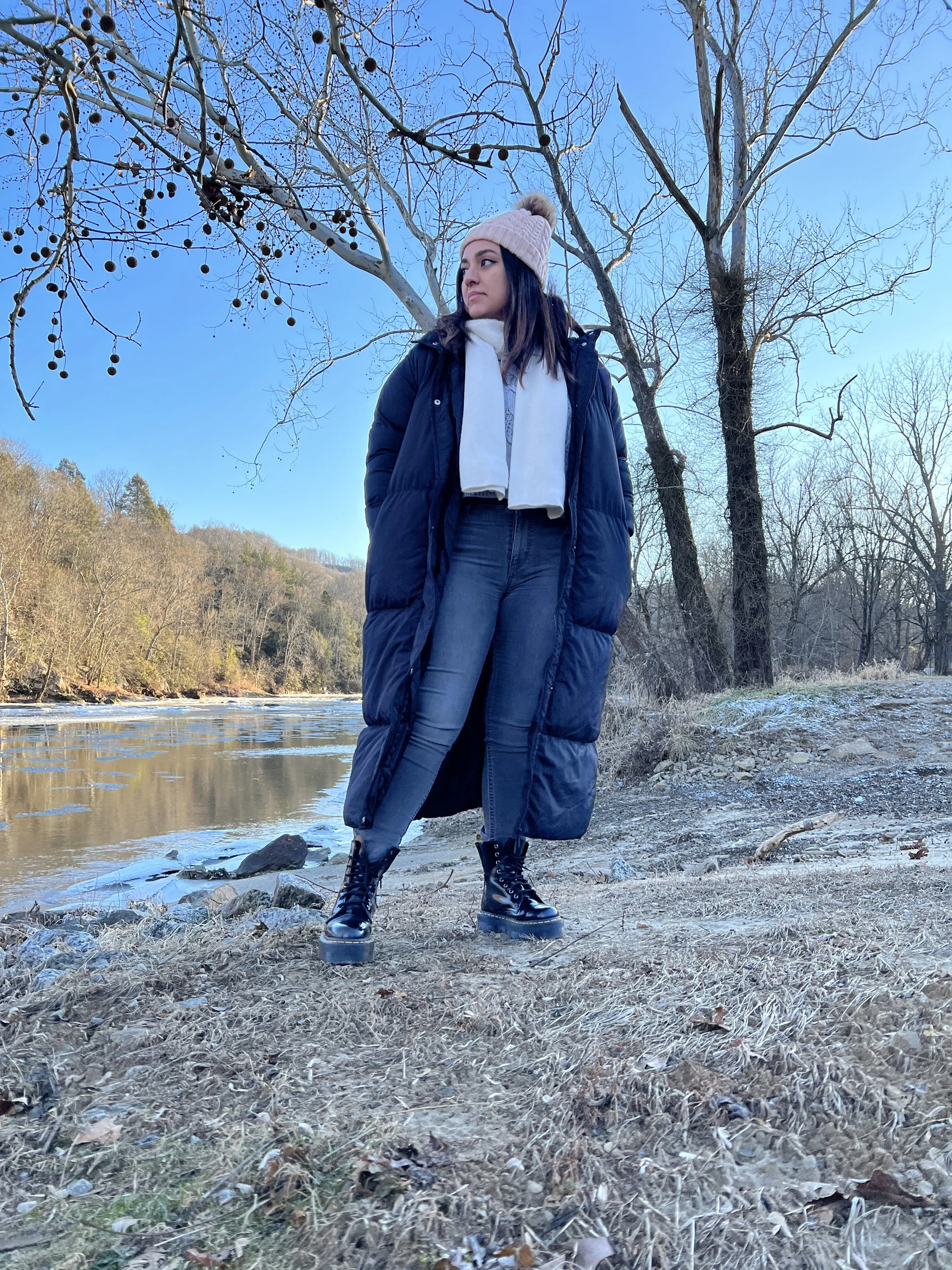 Young woman standing on grassy riverbank in winter clothing, wearing black boots, a navy puffer coat, a beige knit hat with pom-pom, and a white scarf, with bare trees and a river in the background on a clear day.