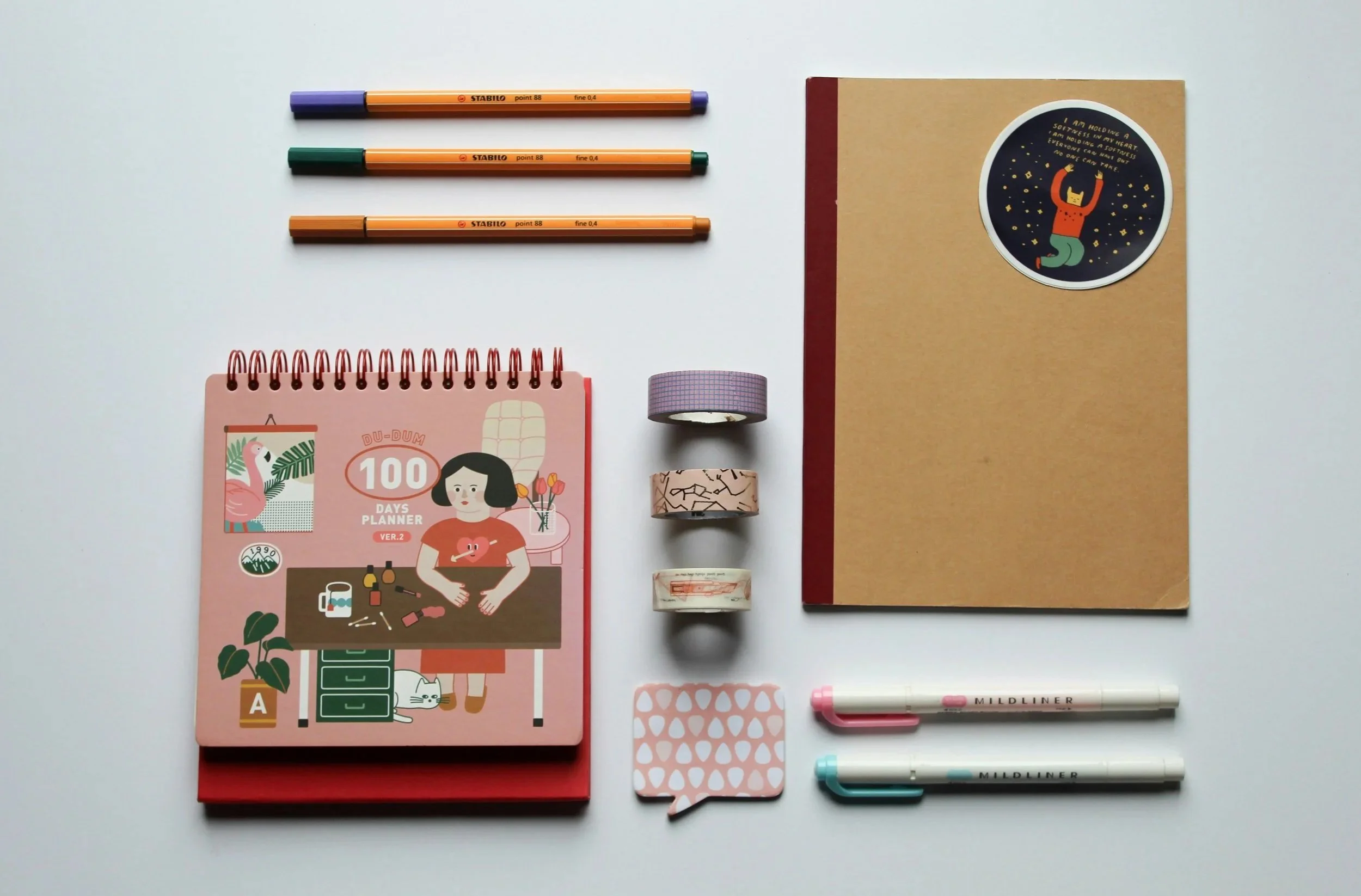 Arrangement of stationery items on a white surface, including three Stabilo markers, a pink 100 days planner, three rolls of decorative tape, a pink speech bubble sticky note, two Mildliner highlighters, a beige notebook with a circle sticker on the cover, and a small sticker sheet.