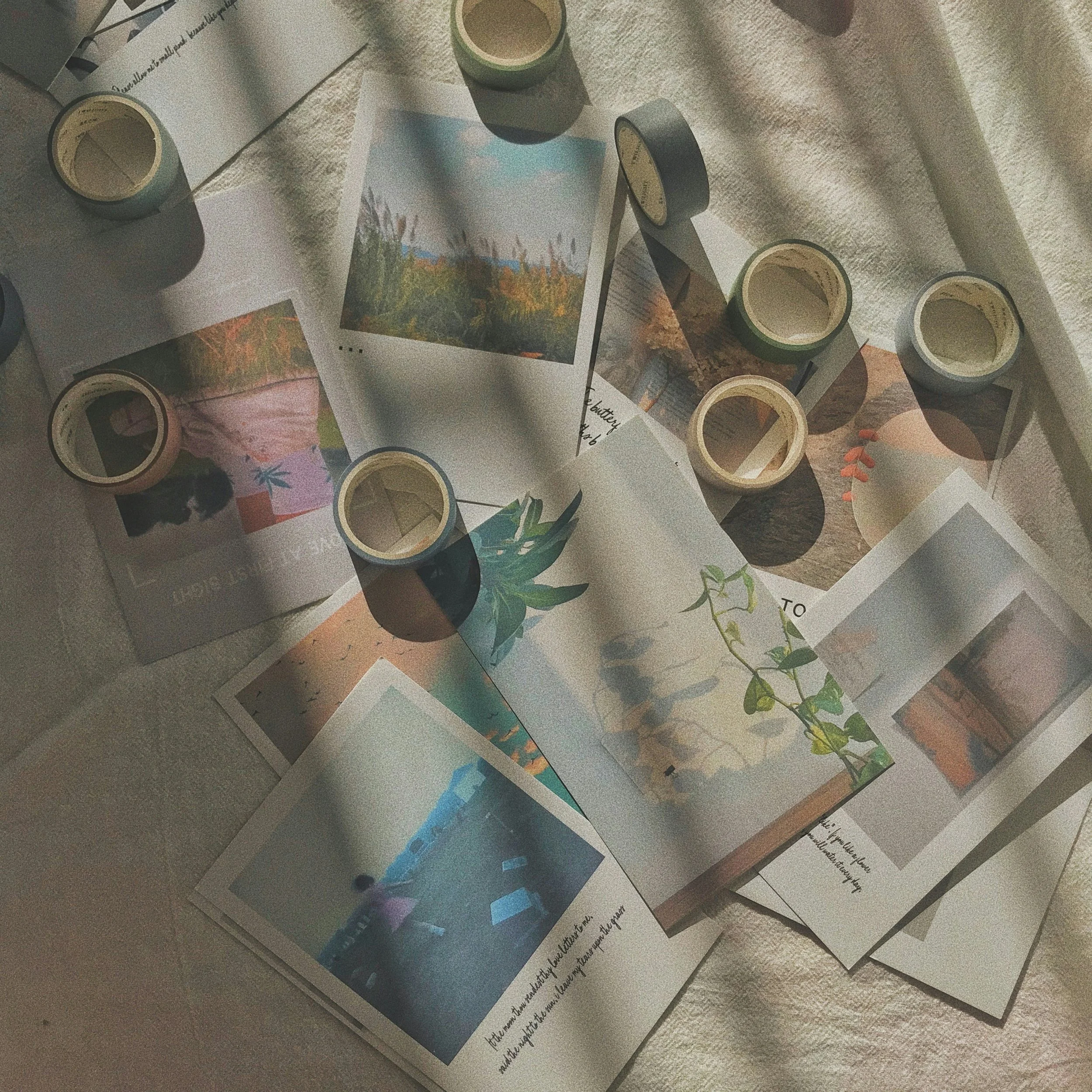 A collage of photographs and small rolls of washi tape arranged on a soft fabric surface, with some photographs showing outdoor scenery and handwritten notes.