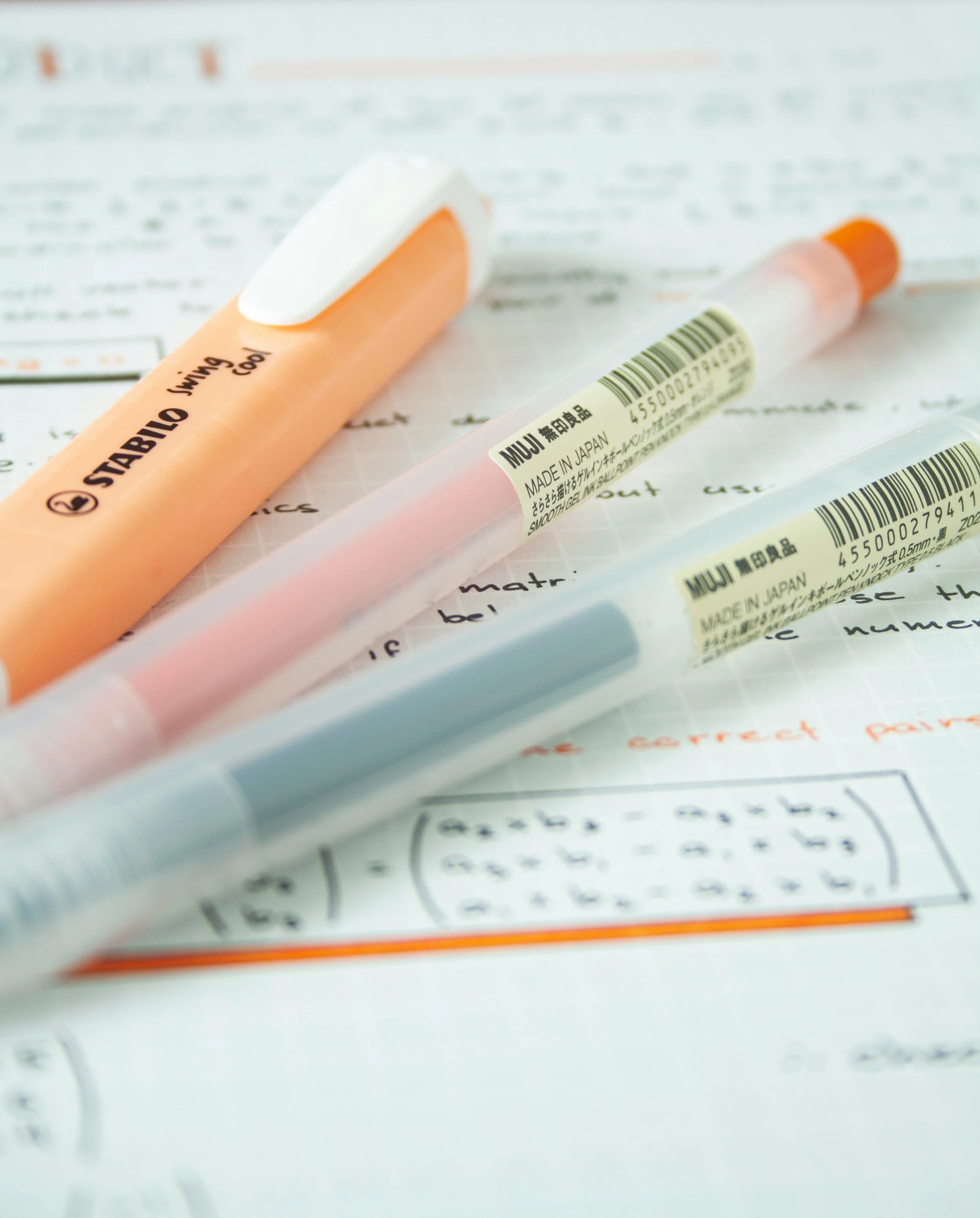 Two pastel-colored Muji pens, one orange and one blue, and one Stabilo highlighter placed on a sheet of paper with handwritten notes and printed mathematics formulas.