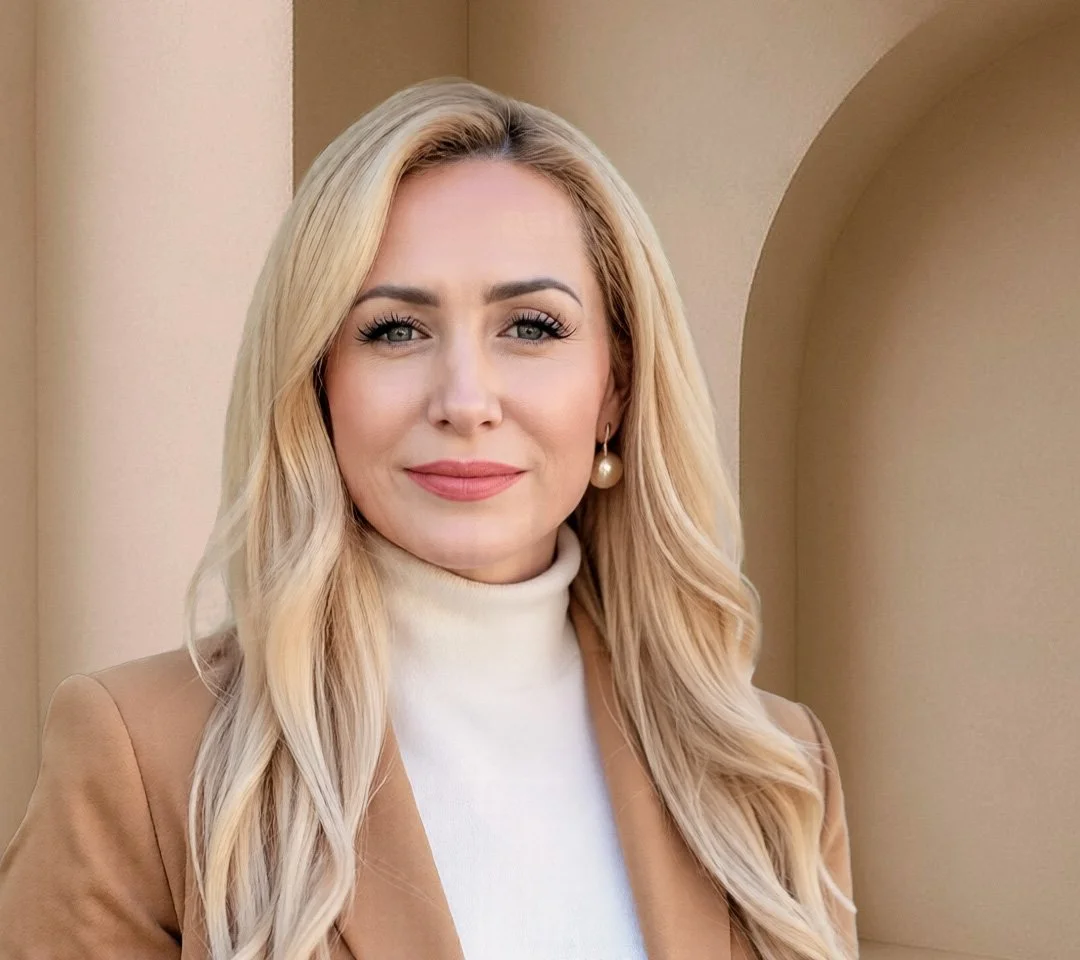 A woman with long blonde hair, wearing a beige blazer over a white turtleneck, and pearl earrings, standing in front of a beige wall and archway.