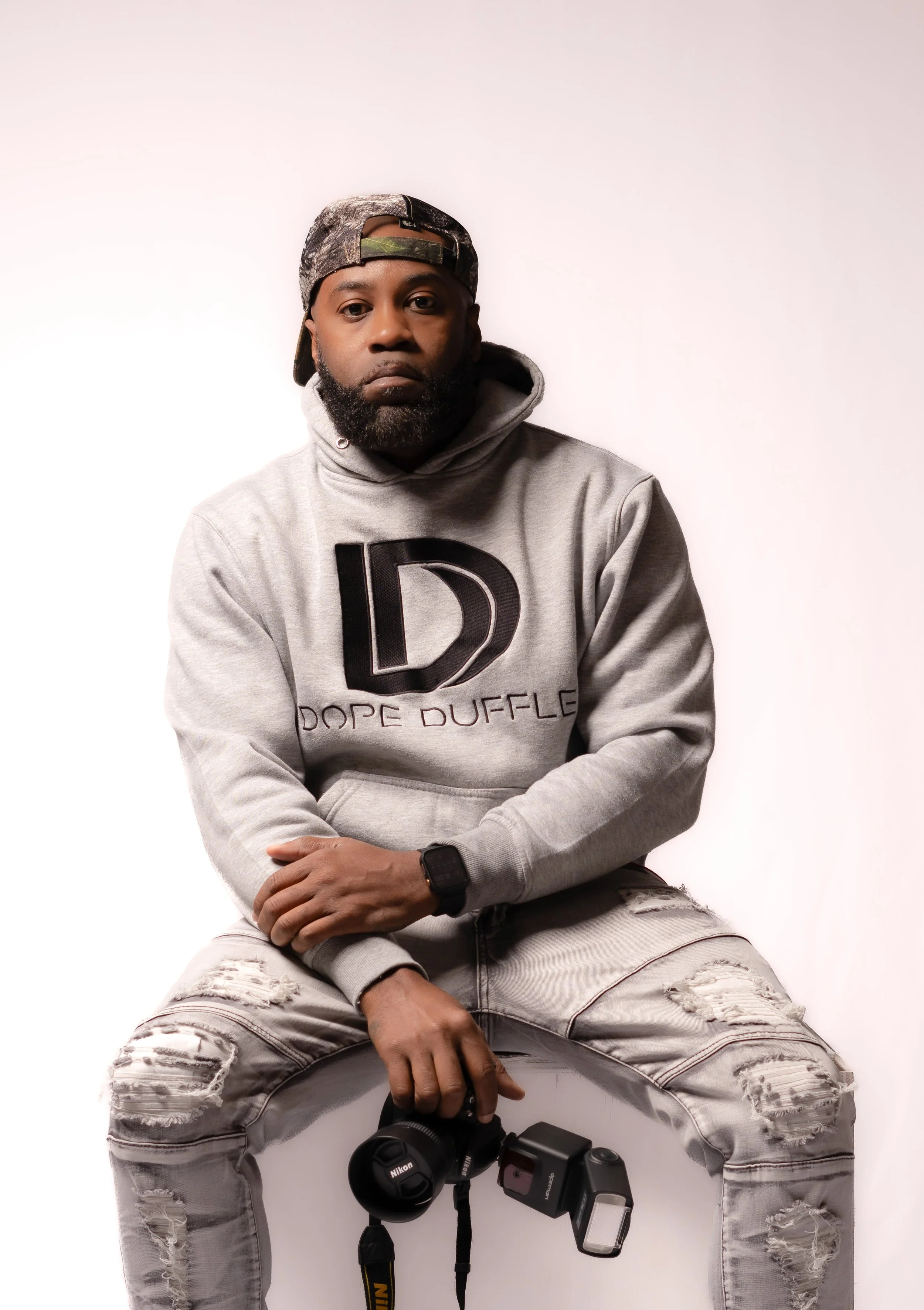Man sitting against a plain white background, wearing a gray hoodie with a large black letter D and the words 'DOPE DUFFLE,' ripped jeans, a camo-patterned baseball cap, and a smartwatch, holding a Nikon camera and an external flash.