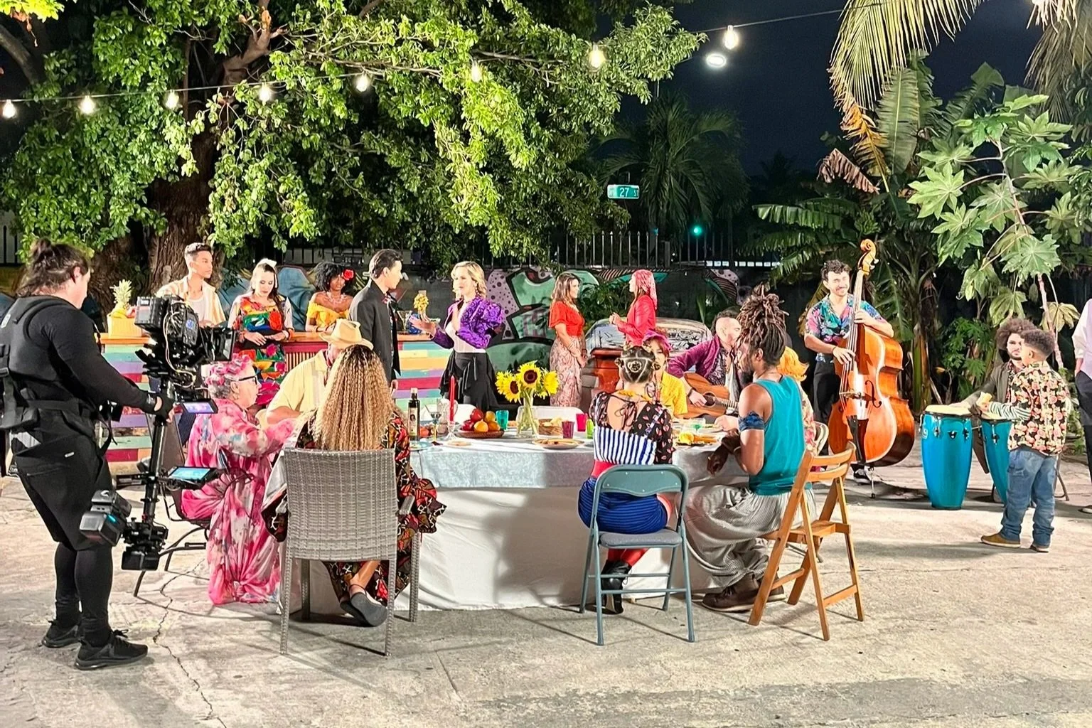 A lively outdoor dinner scene at night with a diverse group of people, some playing musical instruments, others dancing or sitting around a table, under string lights and surrounded by lush greenery.